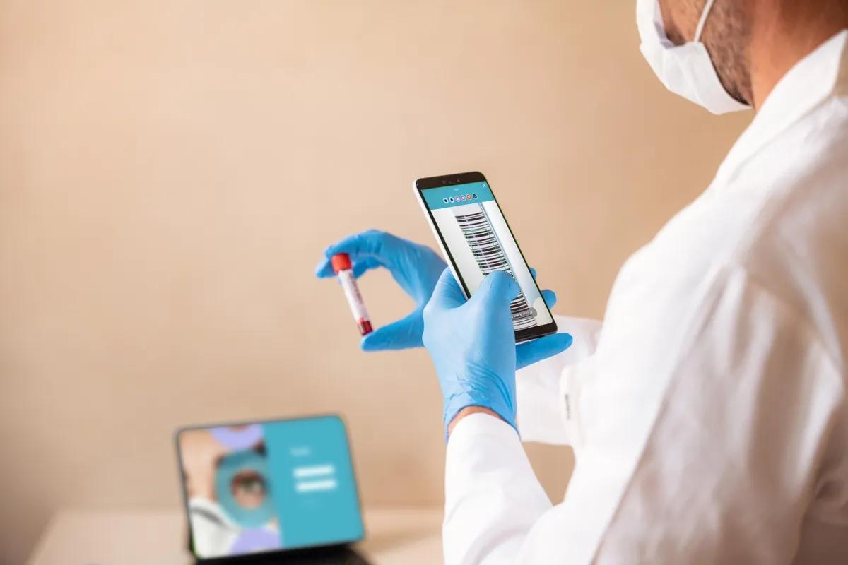 A healthcare professional wearing gloves and a lab coat scans a blood sample with Tracie software on a smartphone, ensuring automated patient-test tube association.