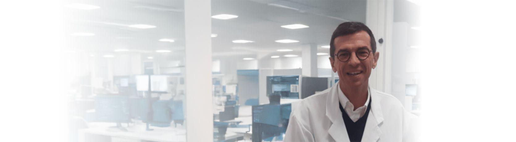 Dr. Christophe Richard Medical Biologist wearing white lab coat standing in a modern laboratory with multiple computer workstations in the background.