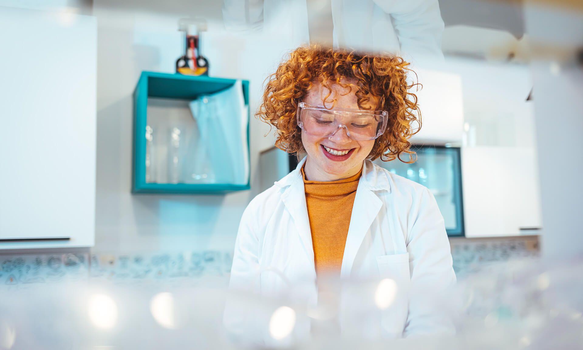 Female lab professional with curly red hair smiling as she works with scientific samples in a brightly lit, modern laboratory.