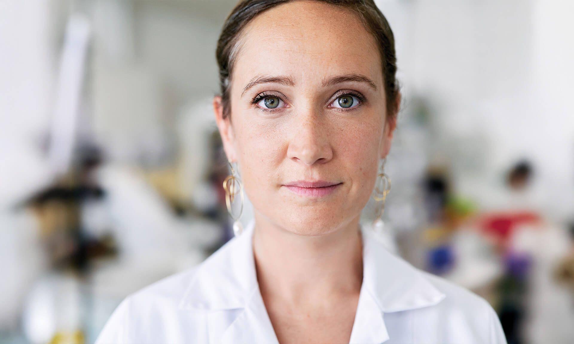 Close-up portrait of a female scientist in a white lab coat, standing in a laboratory environment. She has a focused expression and is wearing subtle gold hoop earrings.