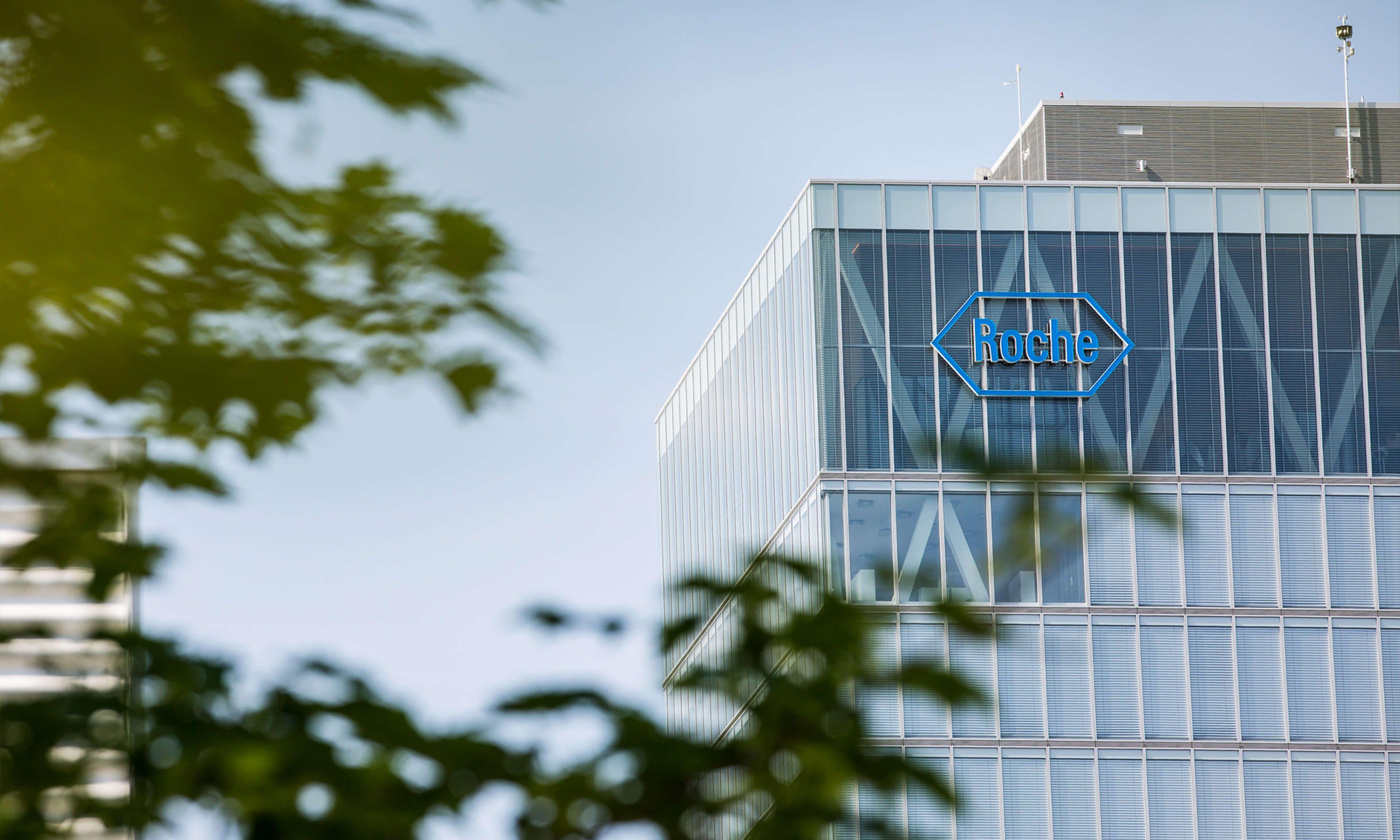 A modern office building with the Roche logo prominently displayed on its glass facade, surrounded by blurred green tree leaves in the foreground.