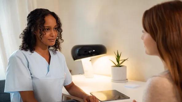 A nurse or medical professional attentively listens to a patient in a consultation room, with a tablet placed on the desk, indicating digital workflow support.