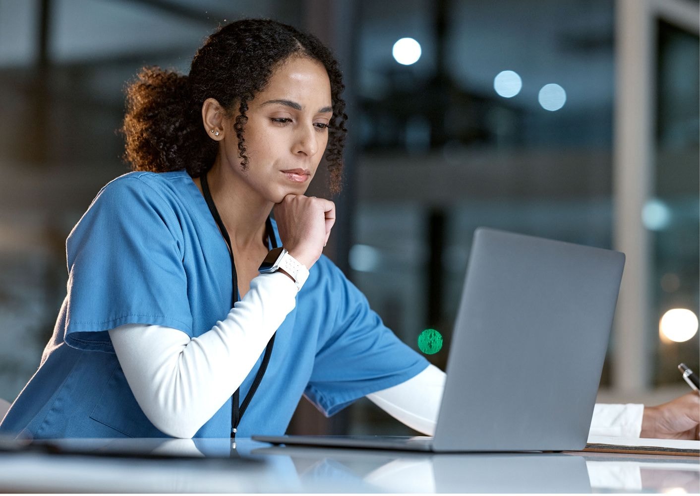  A healthcare professional in blue scrubs sits at a desk, focused on a laptop screen. 