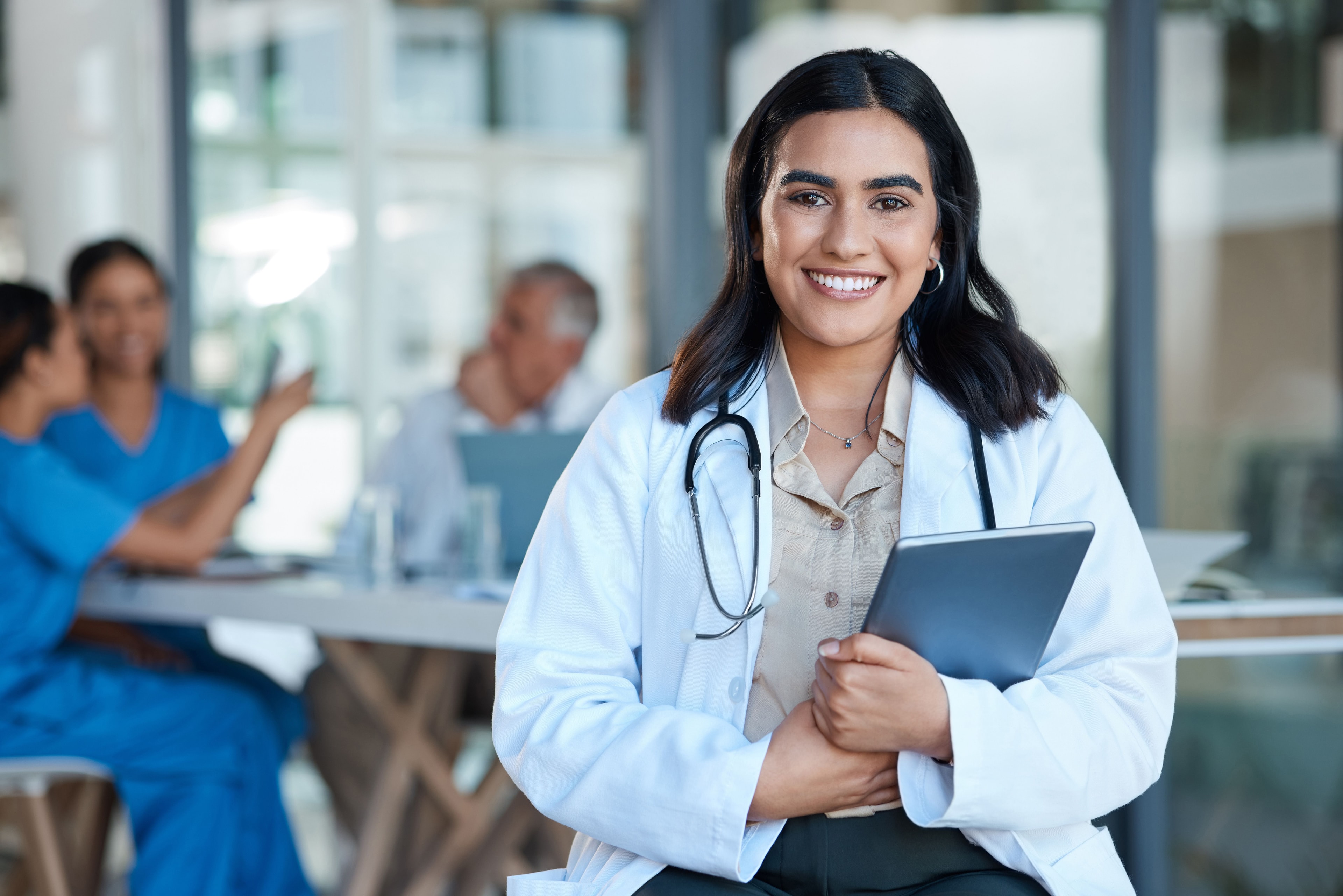A confident female doctor wearing a white coat and stethoscope smiles while holding a tablet in a modern medical setting with colleagues in the background.