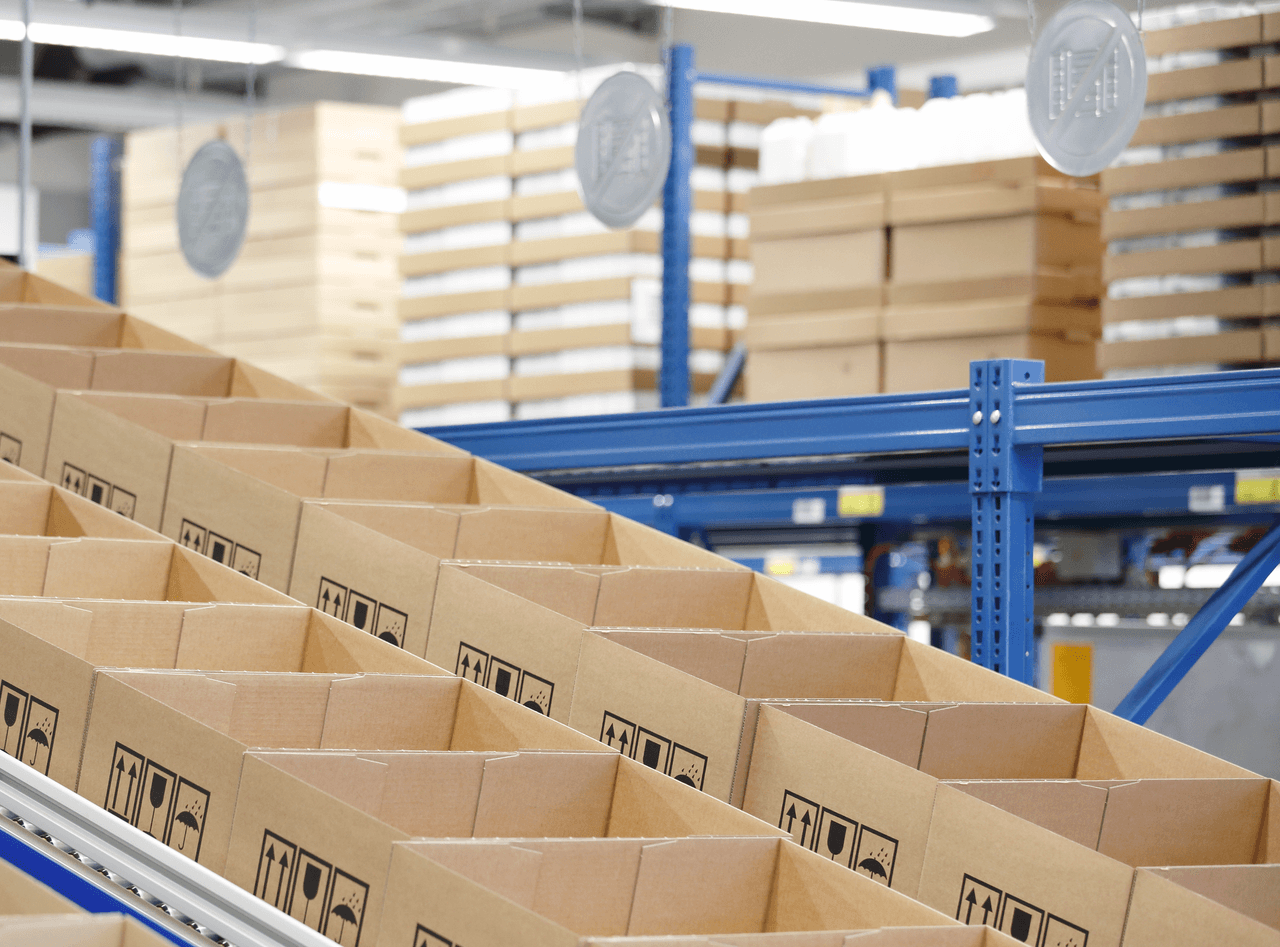 Rows of open cardboard boxes on a conveyor system in a warehouse with shelves of packaged goods in the background.