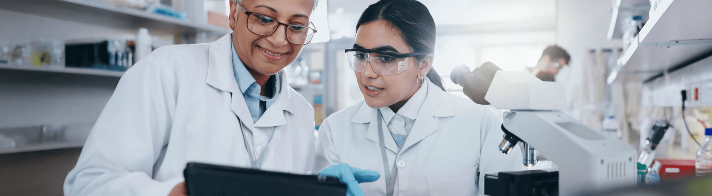 Two scientists collaborate in a laboratory, reviewing data on a tablet near a microscope.