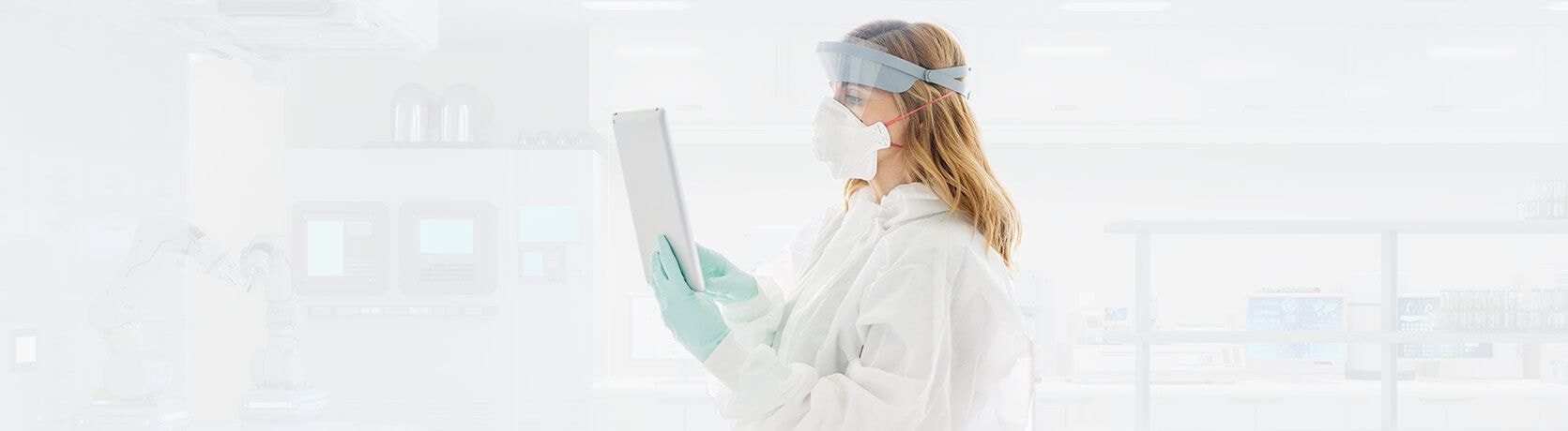 Female lab technician wearing full personal protective equipment (PPE), including gloves, a mask, and a face shield, while using a tablet in a clean, bright laboratory.
