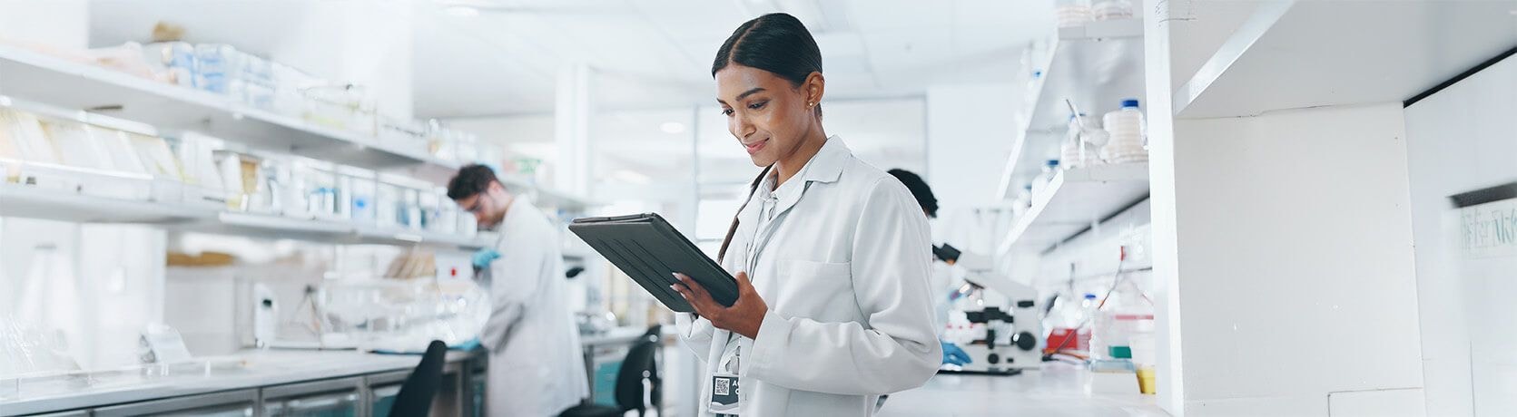 Female lab technician using a tablet in a laboratory with shelves of equipment and supplies in the background.