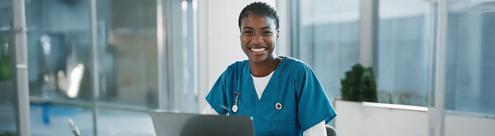 Profesional sanitaria llevando un uniforme azul sentada en un escritorio con una computadora portátil en una consulta médica luminosa.
