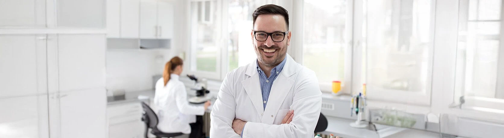 Profesional de laboratorio masculino sonriente que está de pie con actitud de confianza y con los brazos cruzados en un entorno de laboratorio moderno, mientras que otro técnico trabaja en el fondo.
