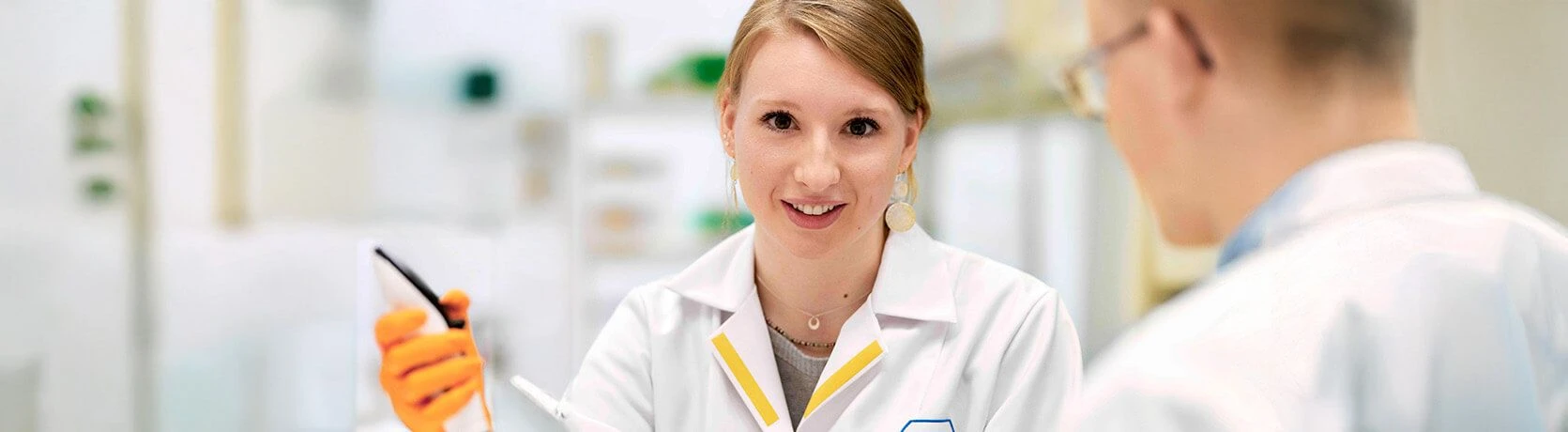 A young female scientist in a lab coat smiling and holding a laboratory instrument while engaged in a discussion with a colleague. The setting is a modern laboratory with bright lighting.