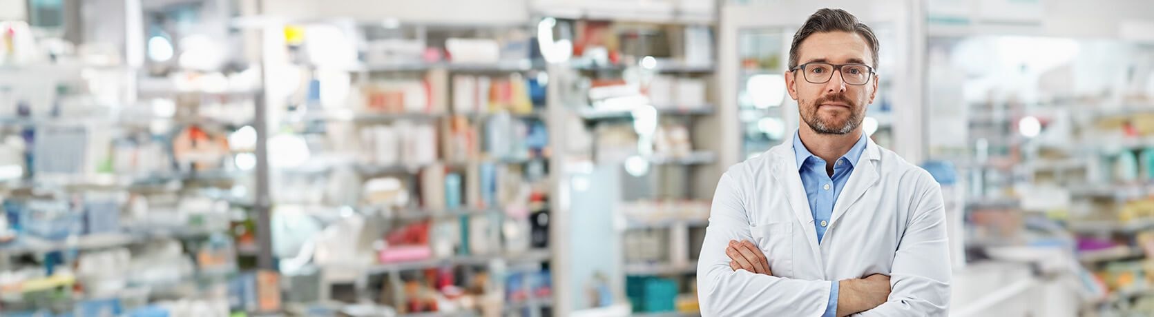Male scientist in a laboratory or pharmacy setting, wearing a white coat and glasses, standing confidently with arms crossed in front of shelves filled with equipment and supplies.