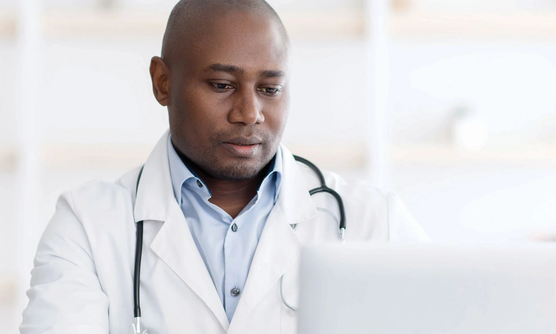 A male doctor wearing scrubs and a lab coat is looking at a computer.