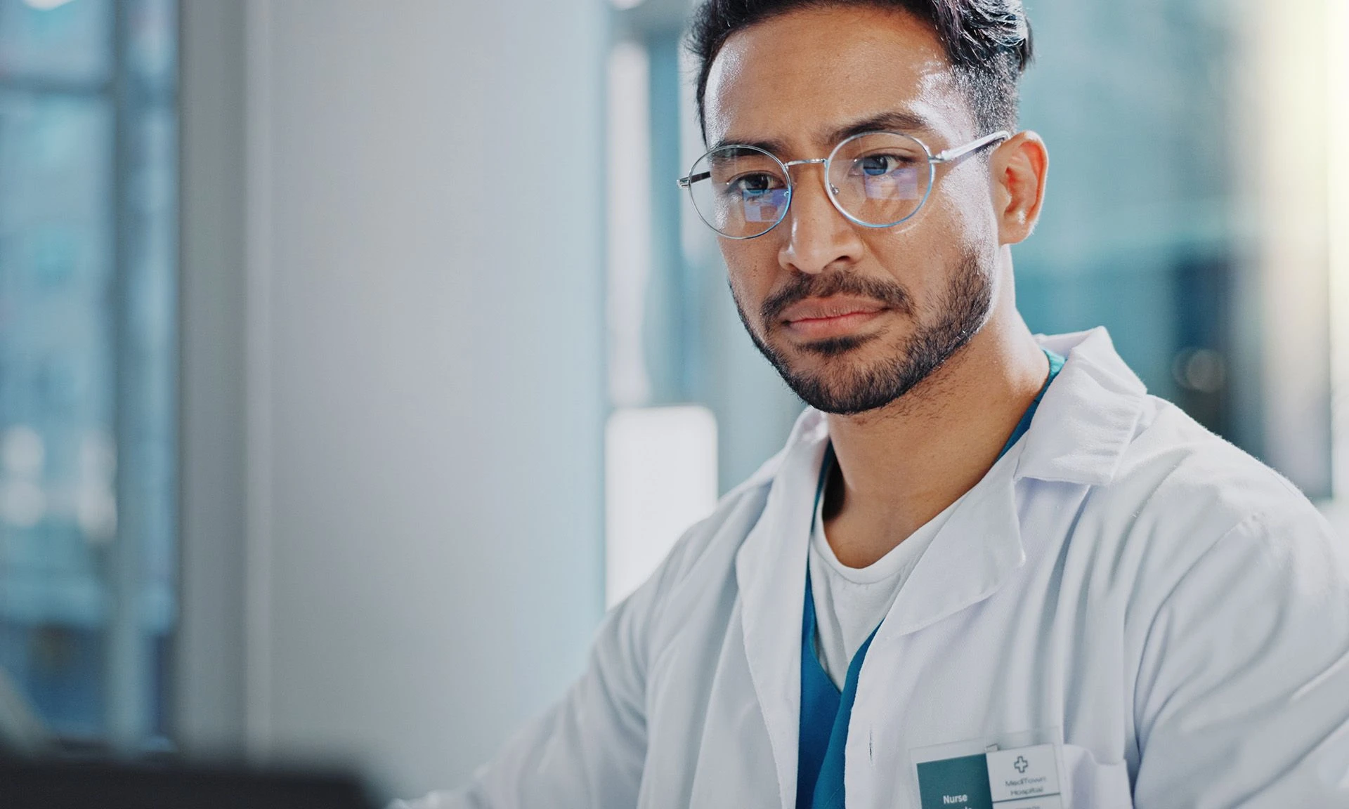 Male healthcare professional in a white coat and glasses, focused on a computer screen in a modern medical facility.
