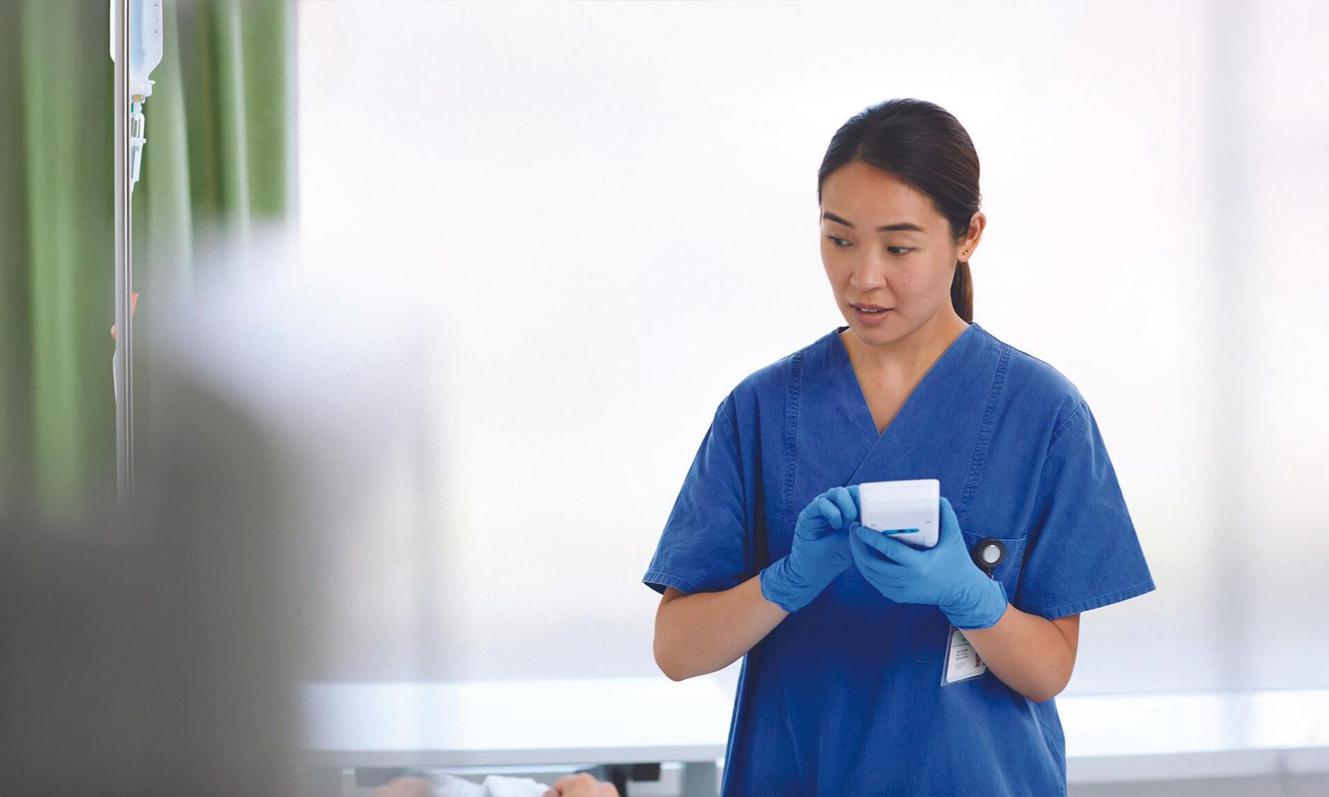 A nurse or technician in blue scrubs and gloves examining a small handheld device in a clinical setting with blurred background elements.