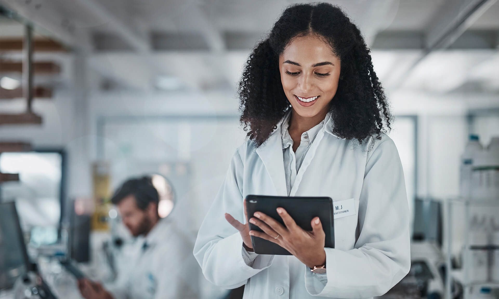 Científica sosteniendo una tableta, sonriendo mientras trabaja en un ajetreado entorno de laboratorio con colegas y equipos avanzados de fondo.