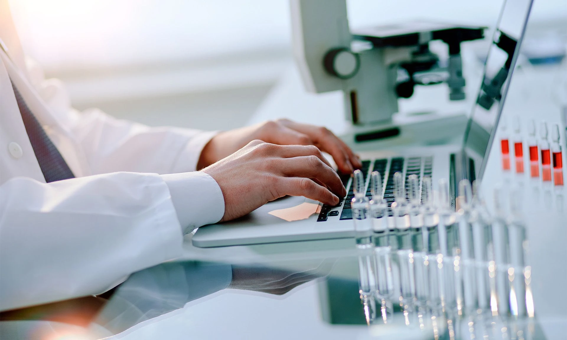 Gros plan des mains d'un scientifique tapant sur un clavier d'ordinateur portable dans un laboratoire, avec des tubes à essai et un microscope en arrière-plan.