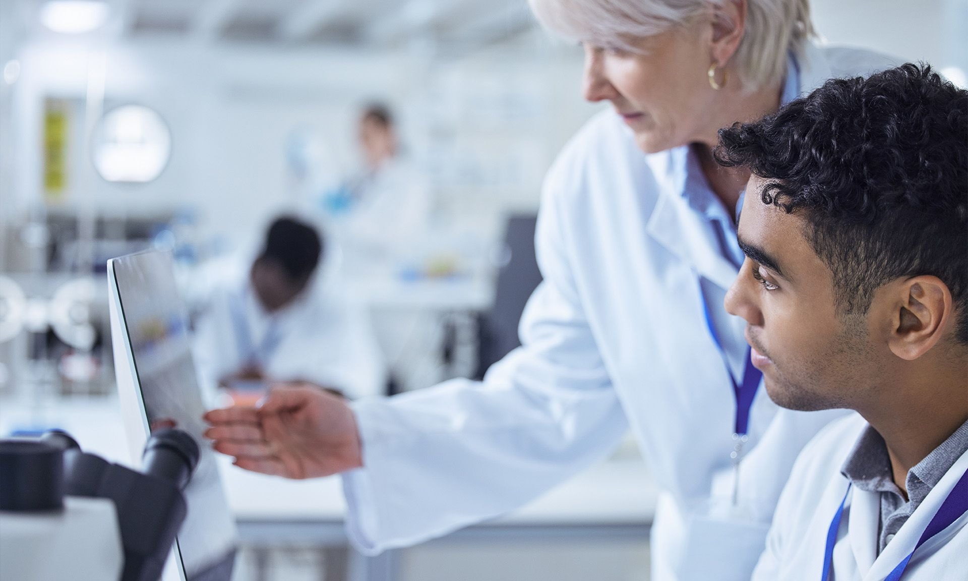 Female healthcare professional guiding a younger colleague in a laboratory setting, both focused on a computer screen.