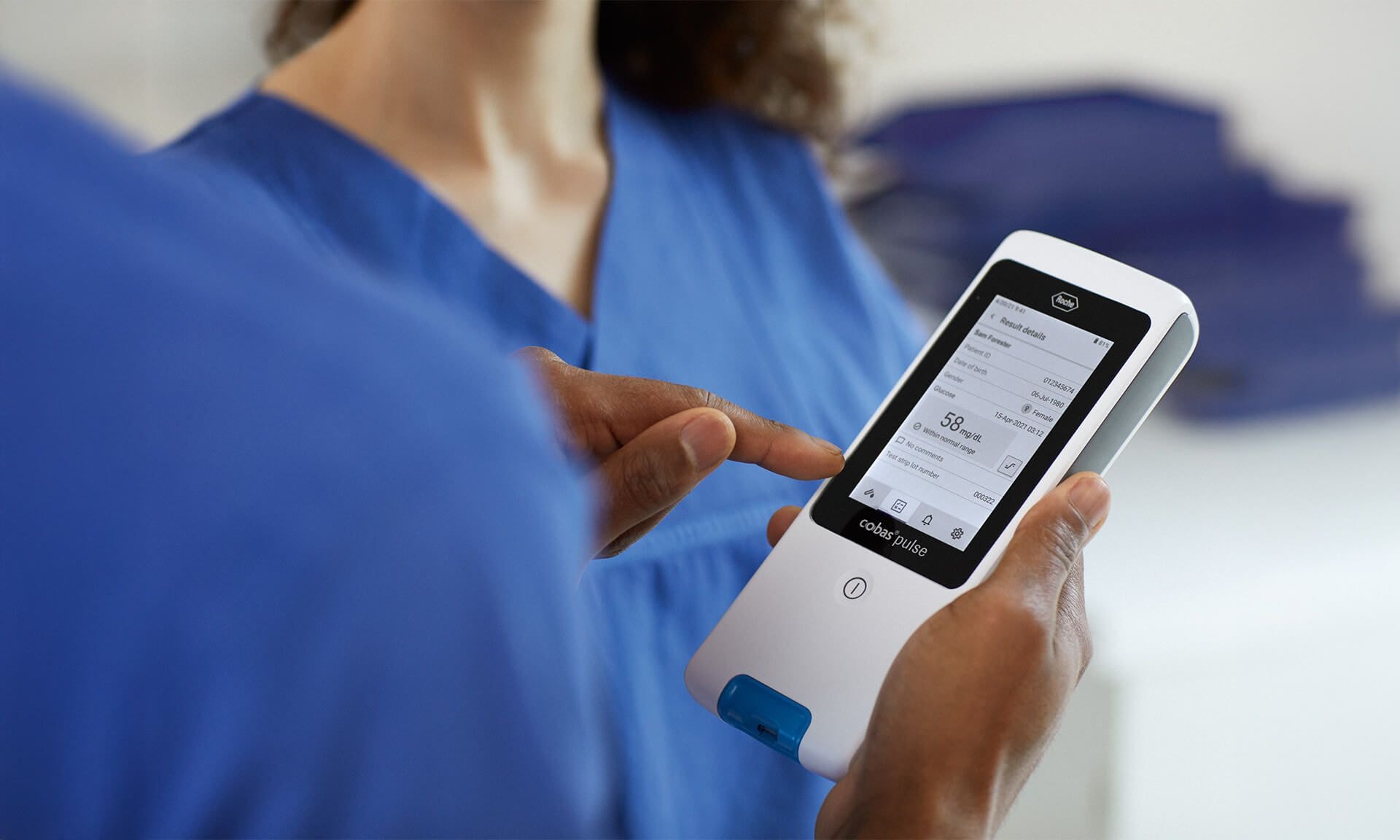 A healthcare professional wearing blue scrubs demonstrating a handheld medical device with a touchscreen interface. The screen displays patient data or diagnostic results.
