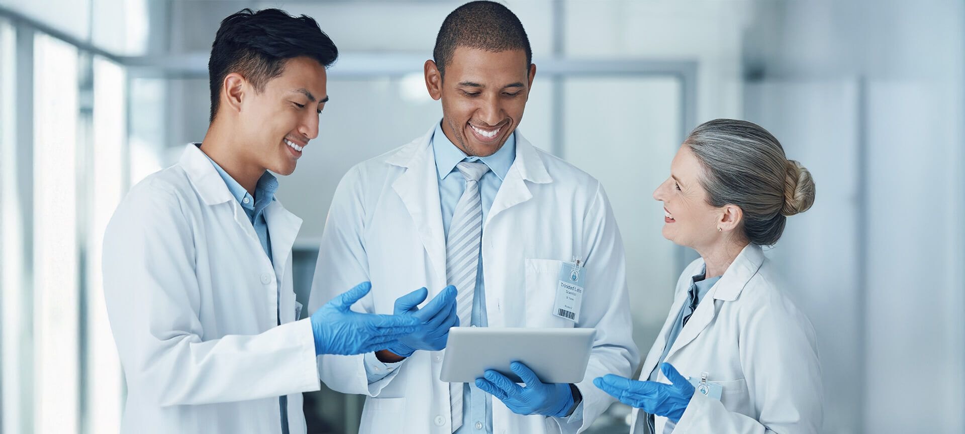 Tre professionisti sanitari in un ambiente clinico, con camici, guanti da laboratorio e tablet digitale, discutono dei dati dei pazienti o di un caso medico.