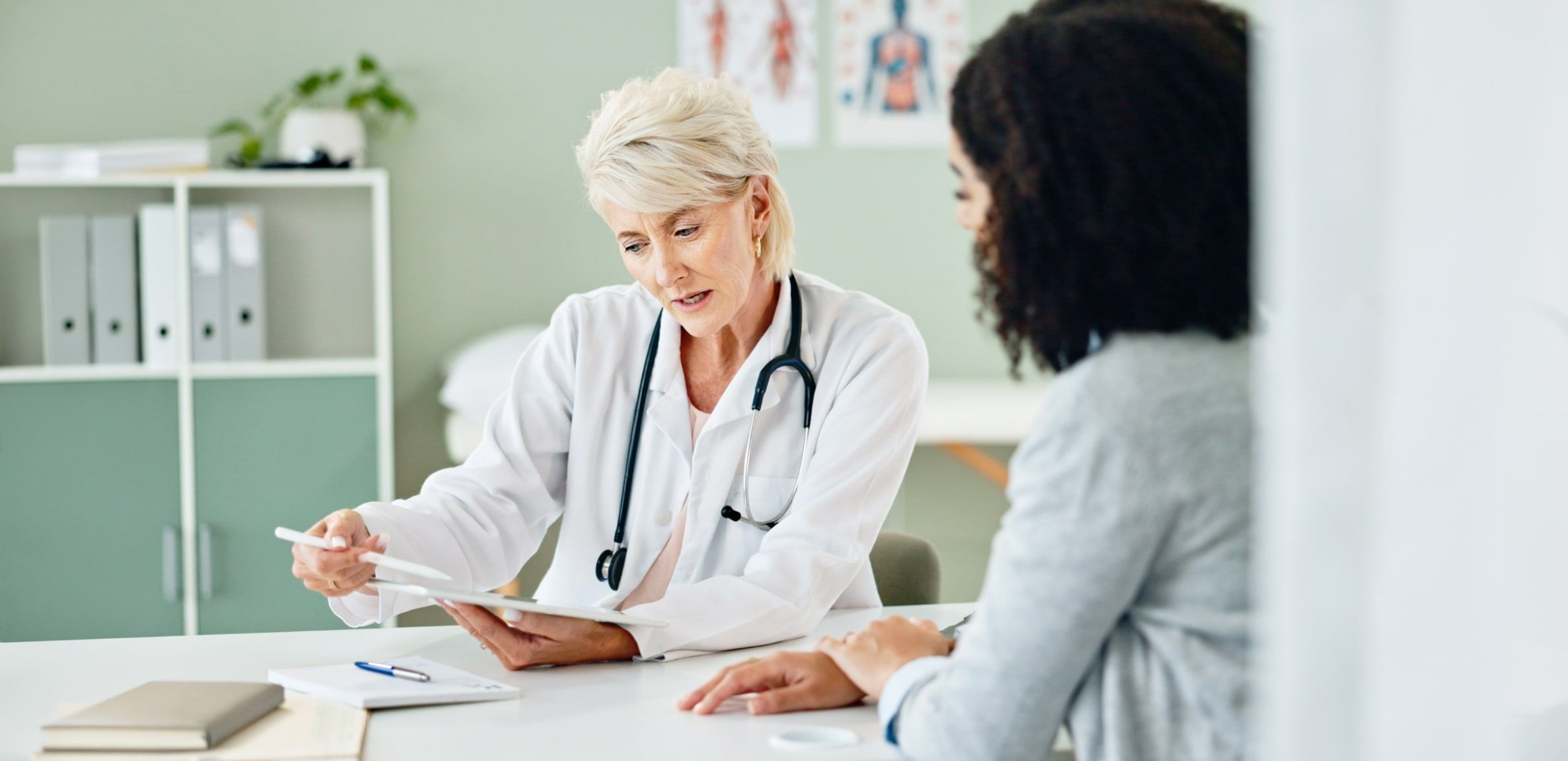 An older female doctor points to charts on a desk during a consultation with a younger female patient.