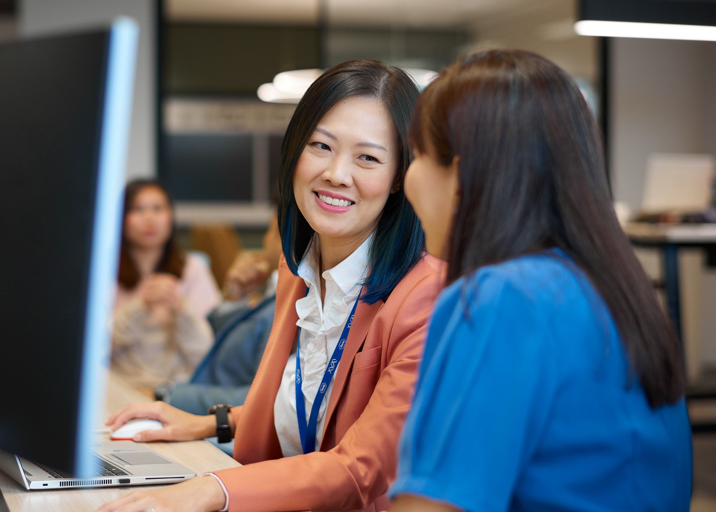 A smiling businesswoman in a coral blazer collaborating with her colleague at a desktop computer.