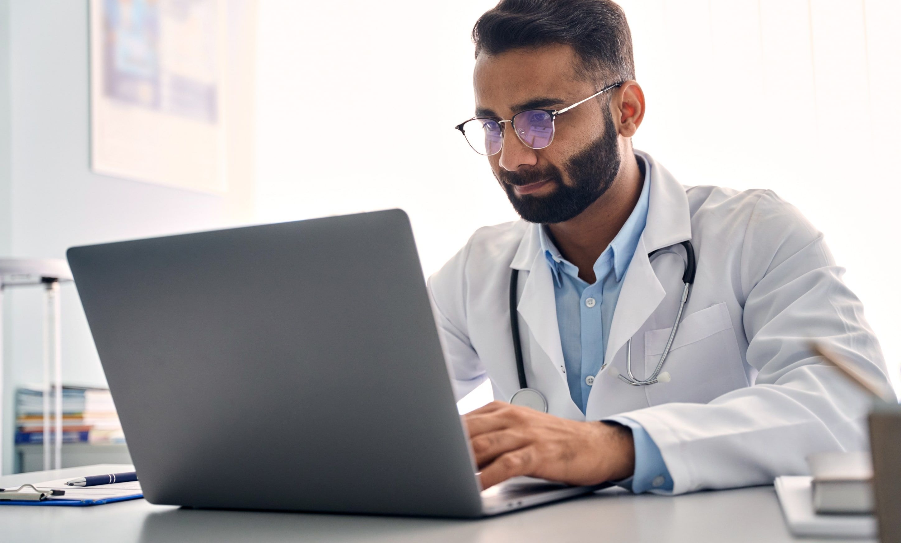 A doctor wearing a white coat works on a laptop at a desk in a medical office.