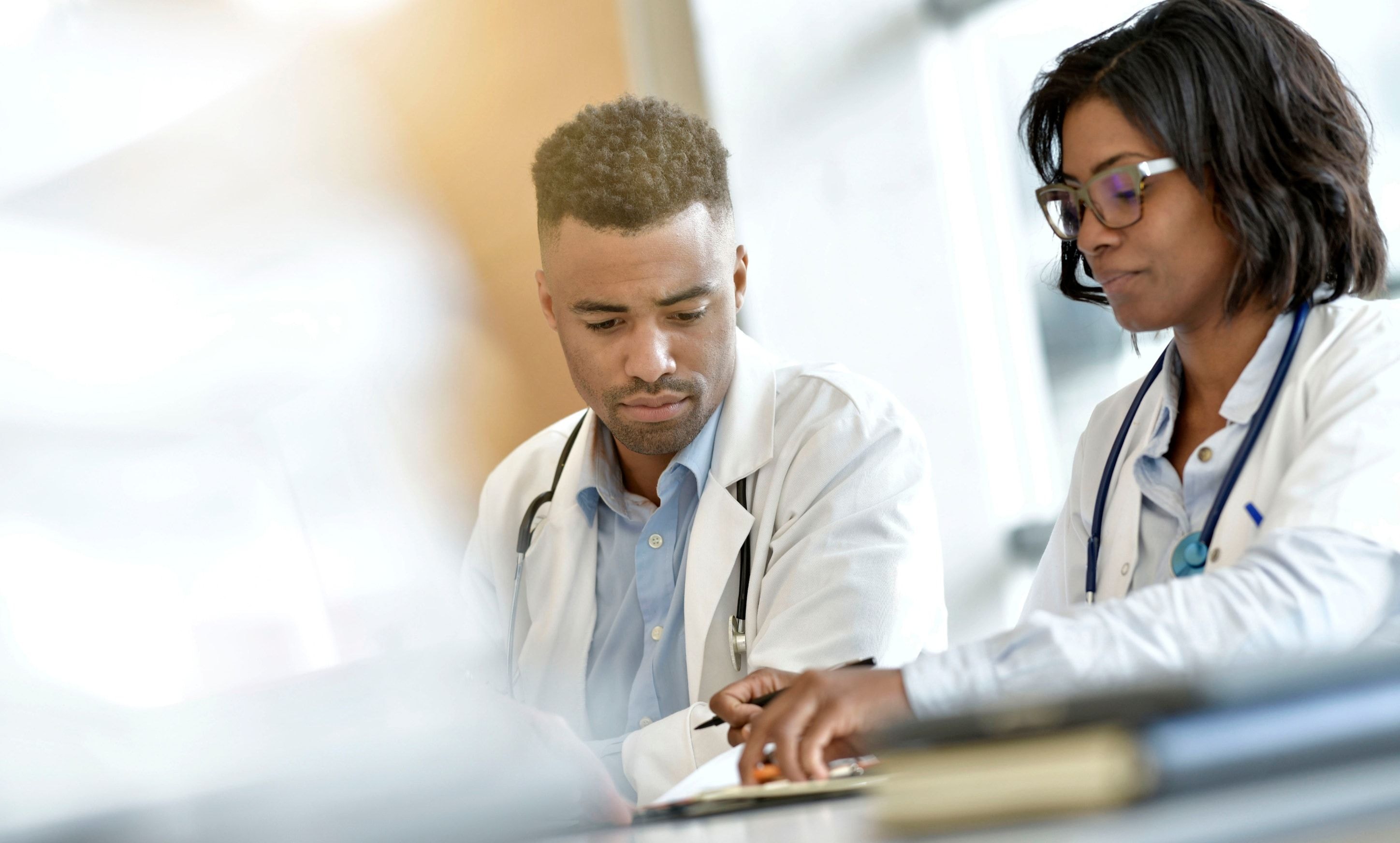 Two healthcare professionals in white coats and stethoscopes reviewing medical charts together at a desk