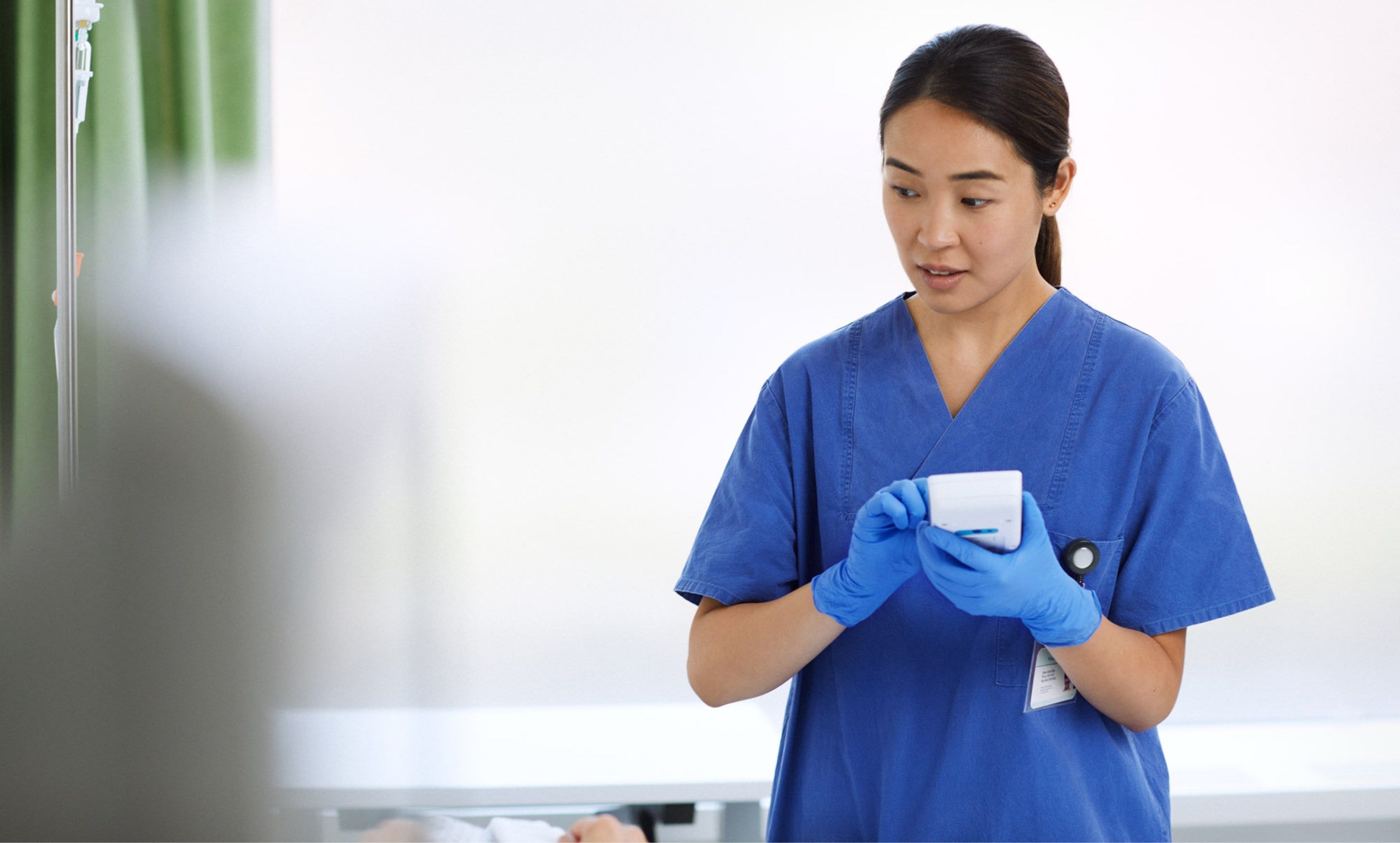 A nurse in blue scrubs and gloves holds a handheld medical device in a hospital setting.