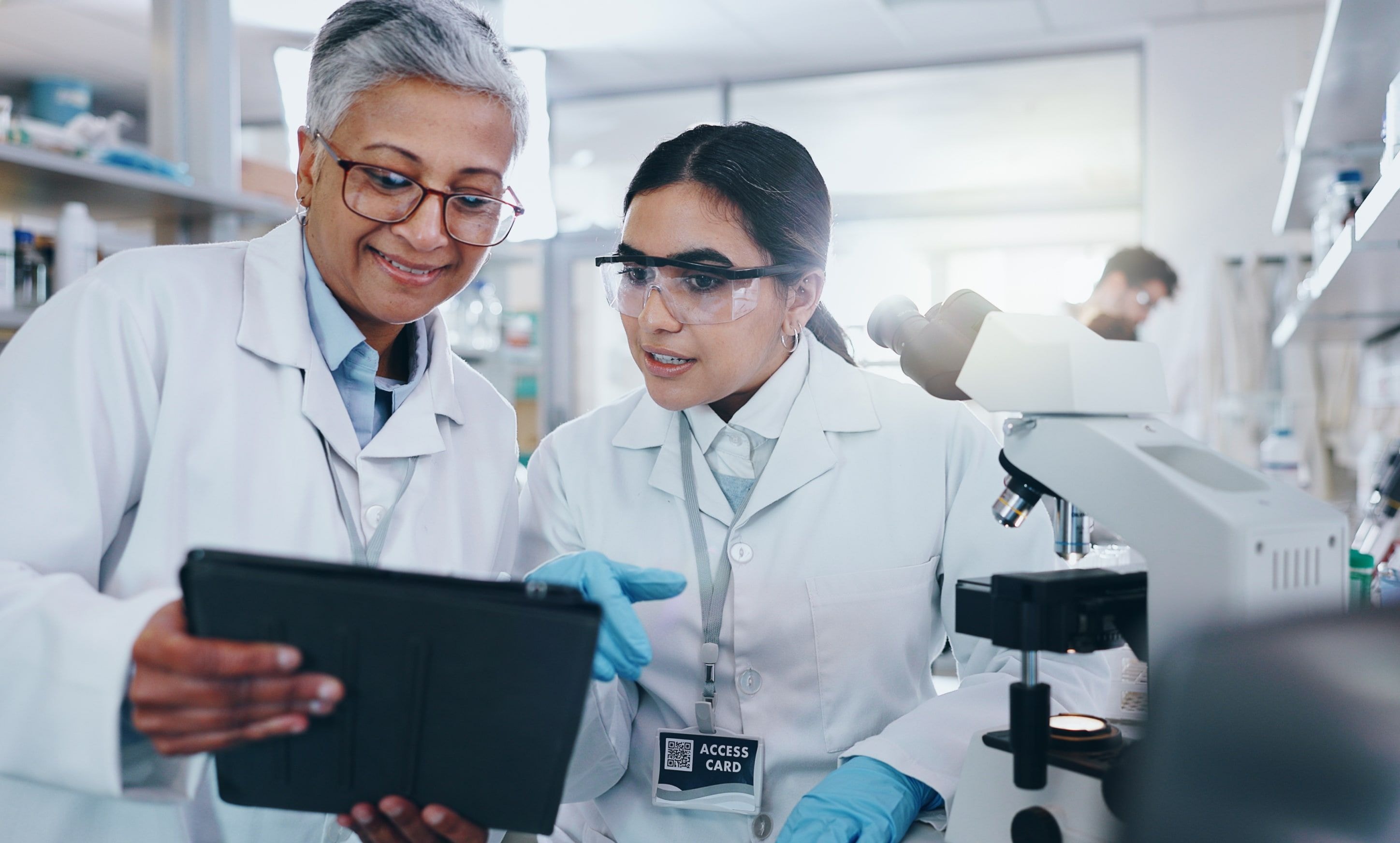 Two scientists in lab coats discuss data on a tablet near a microscope in a laboratory.