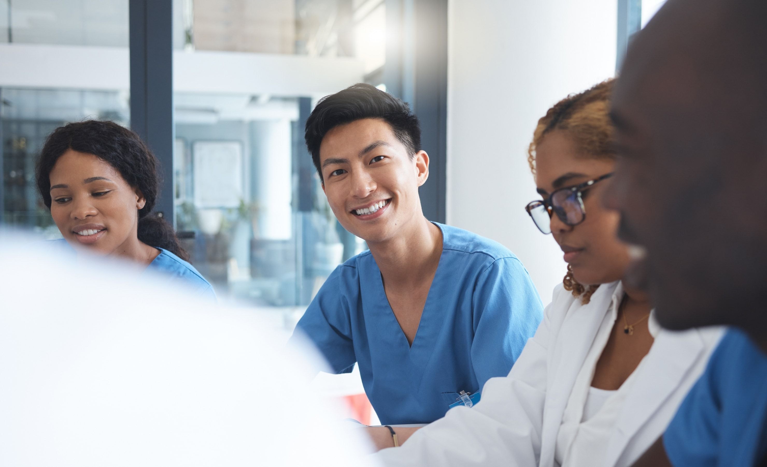 A diverse group of healthcare professionals in blue scrubs and a lab coat having a meeting, with one man smiling.