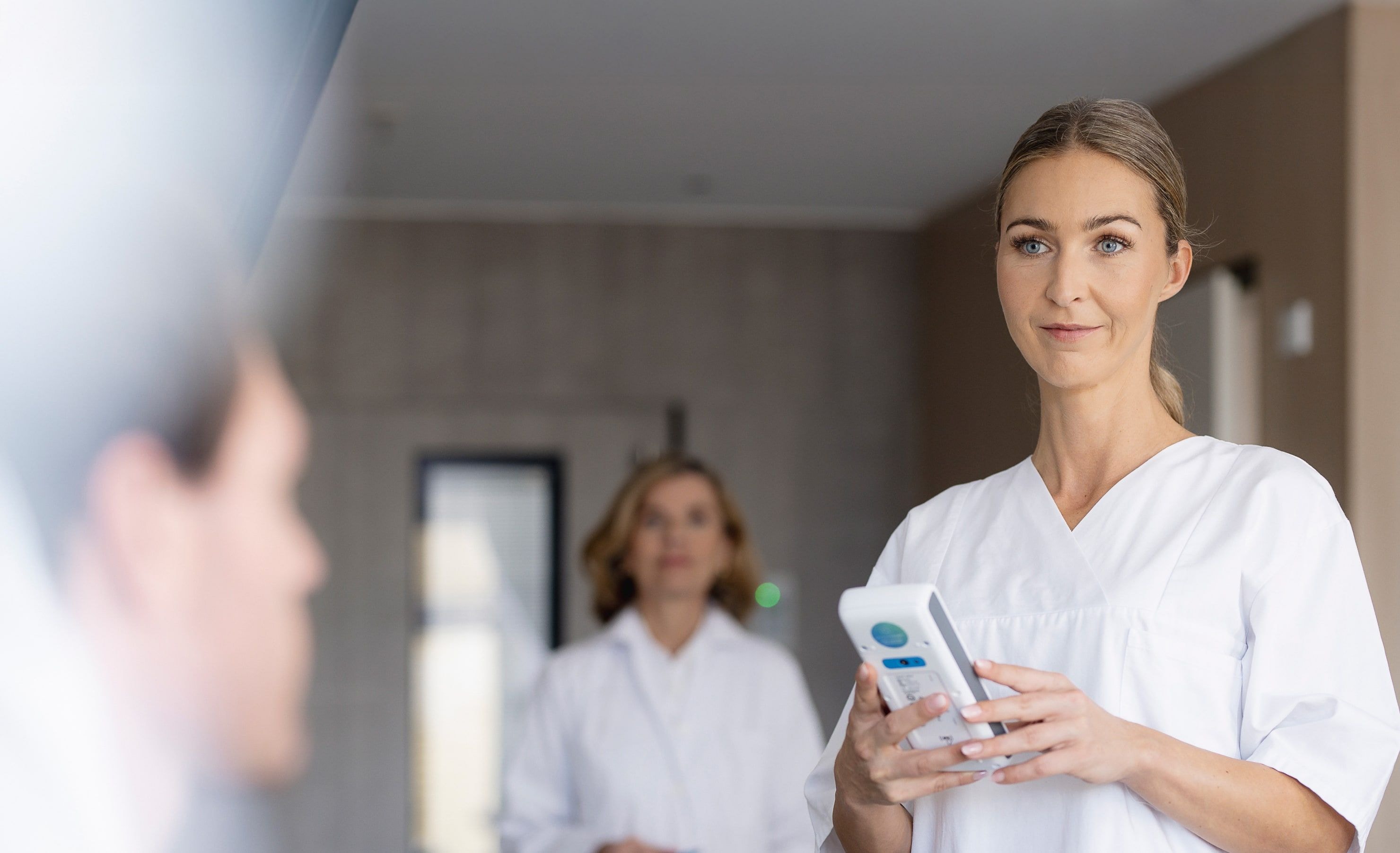 A medical professional in white clothing holds a handheld electronic device indoors.
