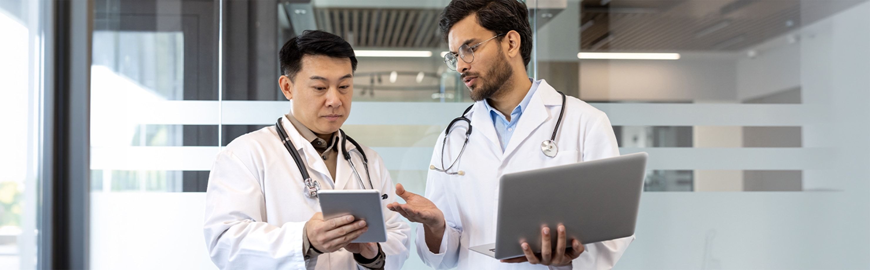 Two doctors in white coats collaborating with a tablet and laptop in a modern medical office.