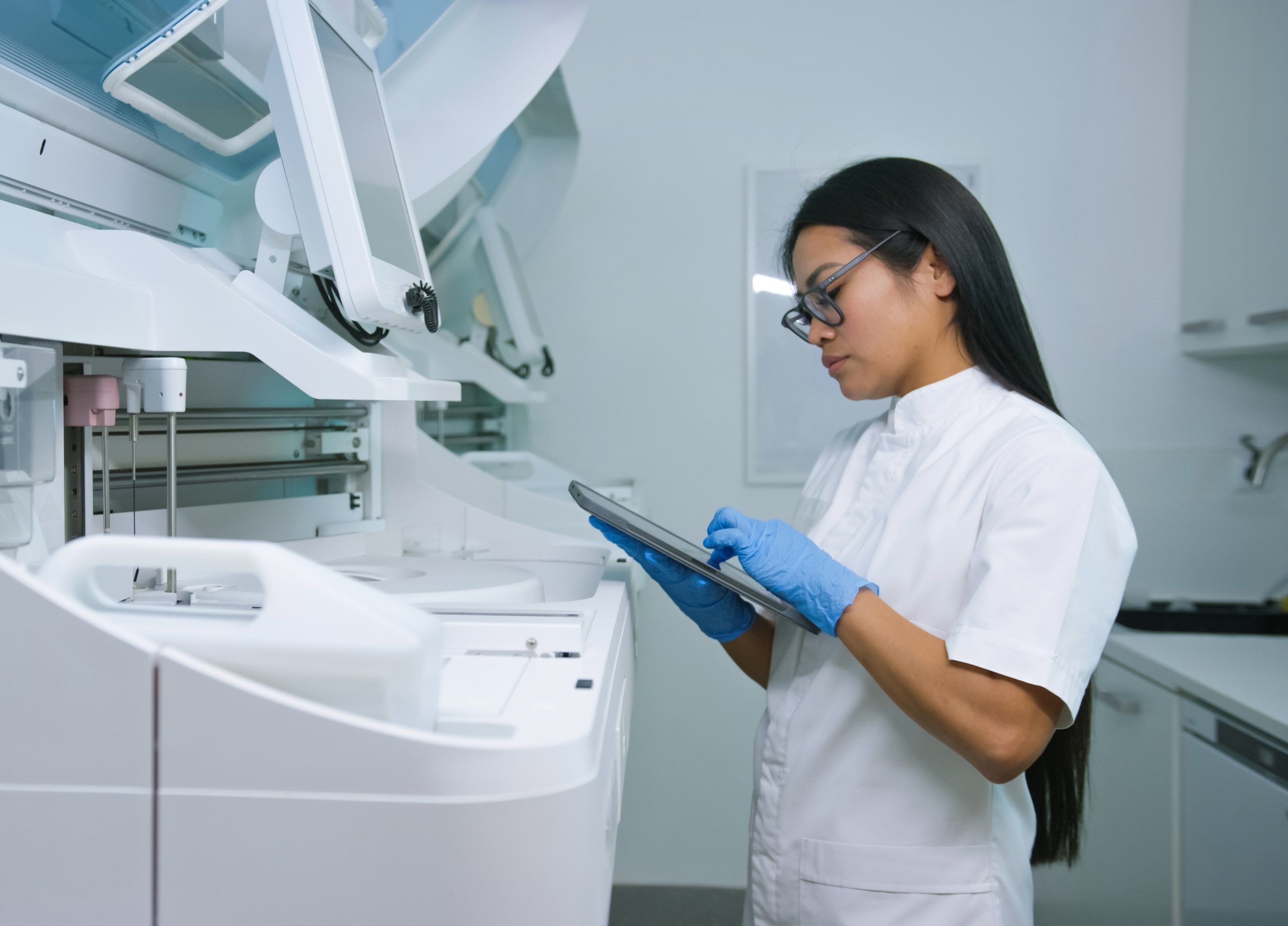 A laboratory technician in gloves operates an advanced medical analyzer using a digital tablet.
