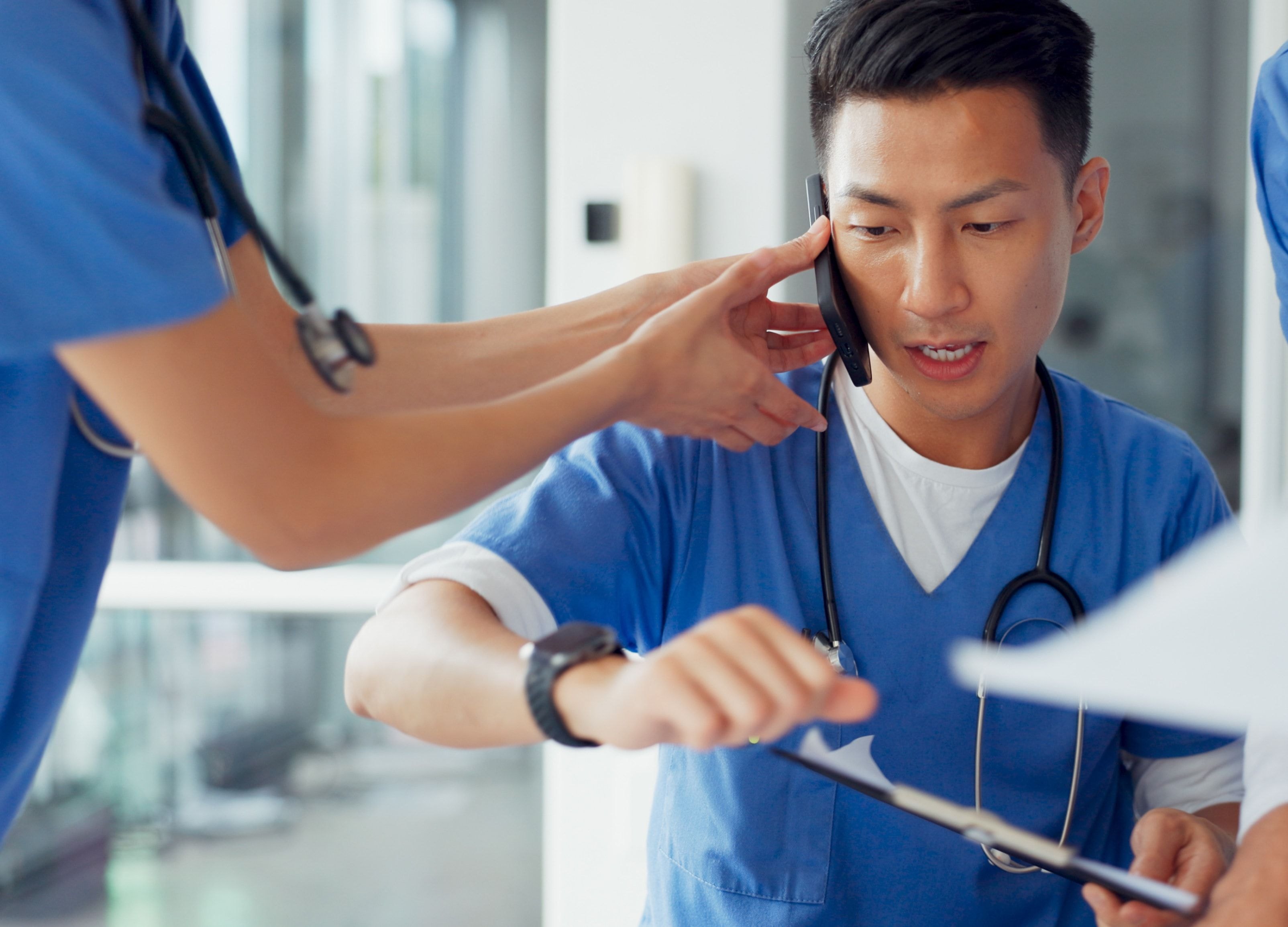 A healthcare worker in blue scrubs holding a clipboard and talking on a phone with a focused expression.