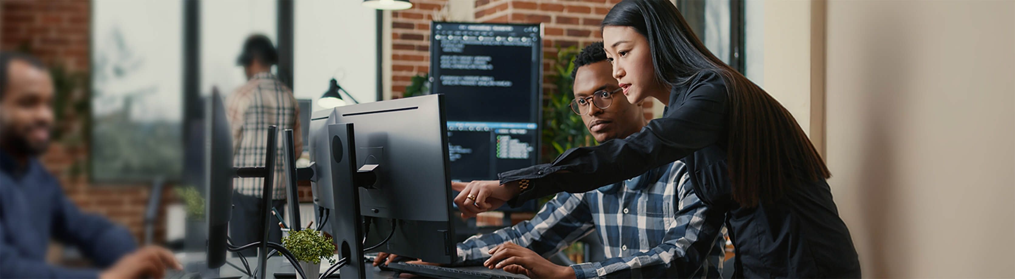 Female IT professional pointing at a monitor, discussing cybersecurity solutions with a colleague in a modern office setting.