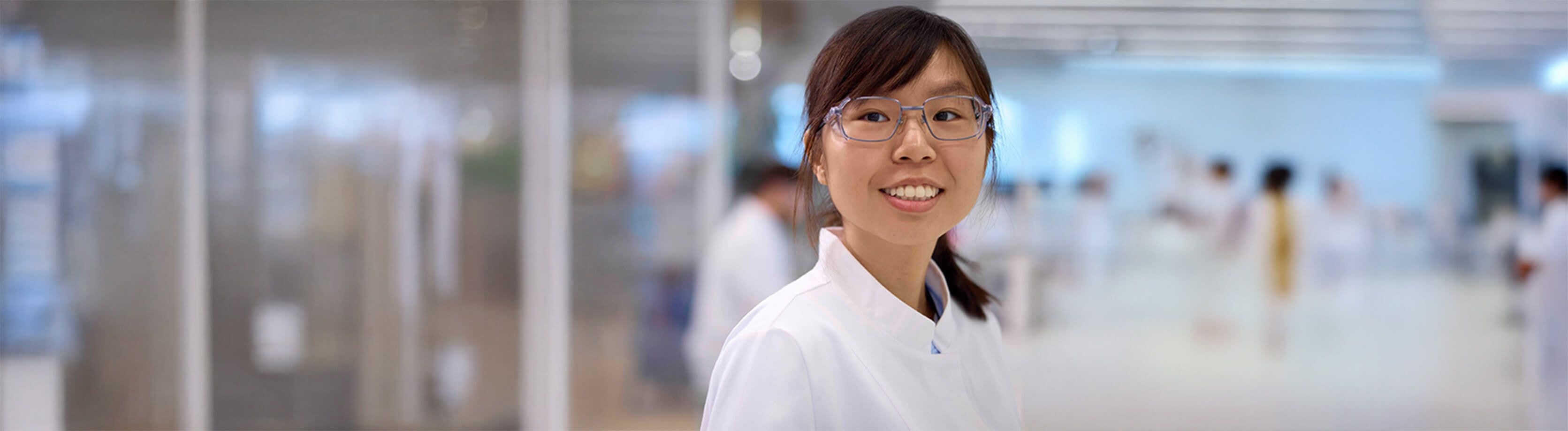 Femme scientifique souriante portant des lunettes et une blouse de laboratoire dans un cadre de laboratoire moderne et lumineux avec des collègues flous en arrière-plan.