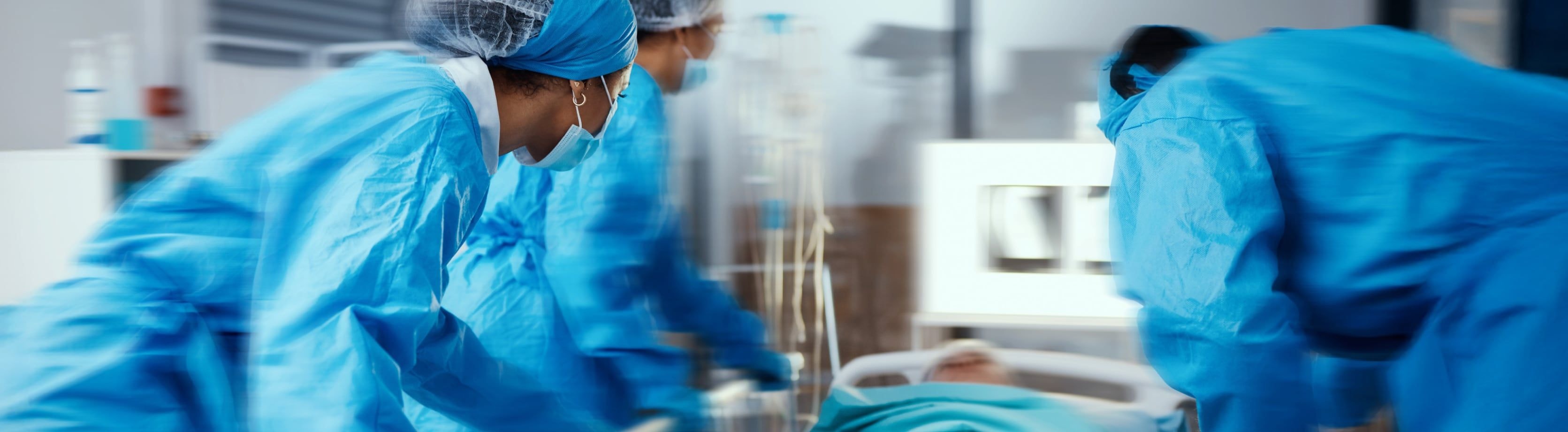 Close-up of medical professionals in blue protective gear attending to a patient in a hospital bed, suggesting an urgent care or surgical setting.