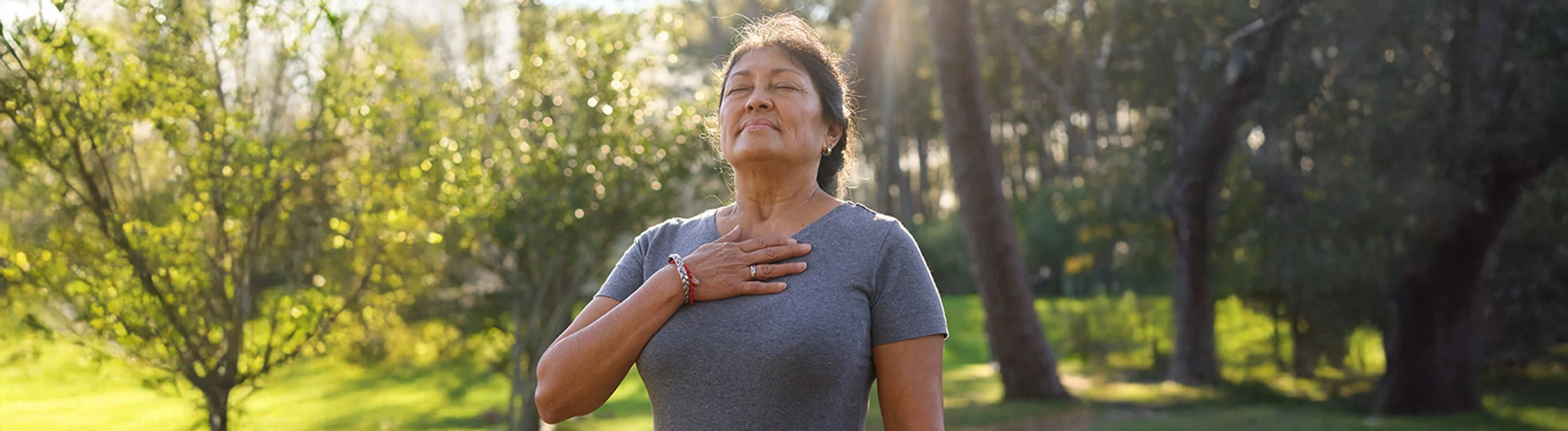 A woman standing outdoors in a peaceful park, closing her eyes with a serene expression and placing her hand on her chest as she breathes deeply