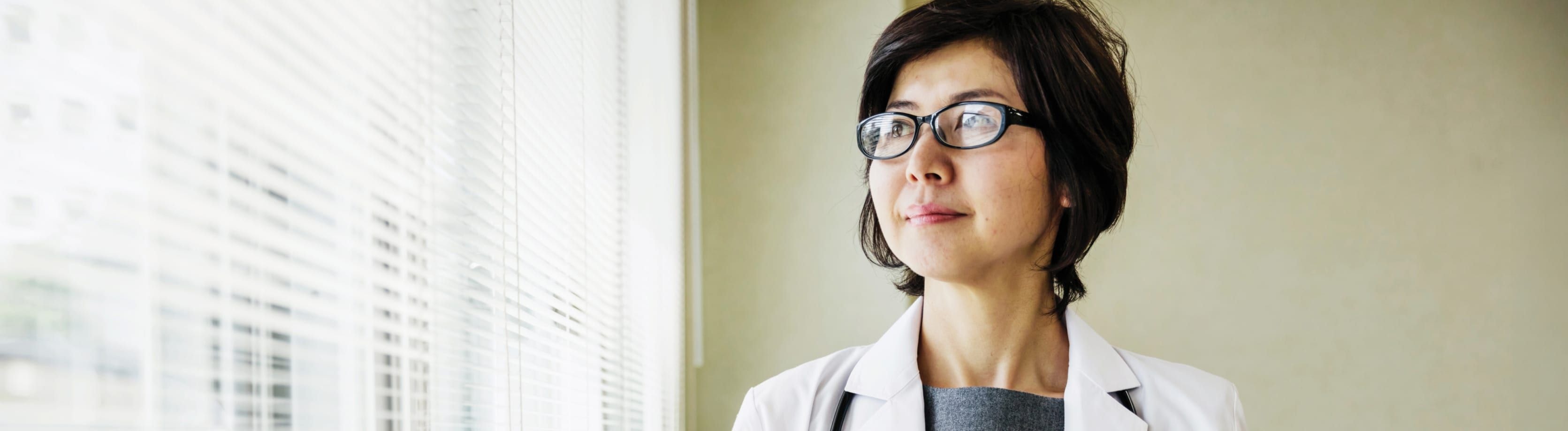 A person in a white lab coat standing near a window with blinds, gazing outward.