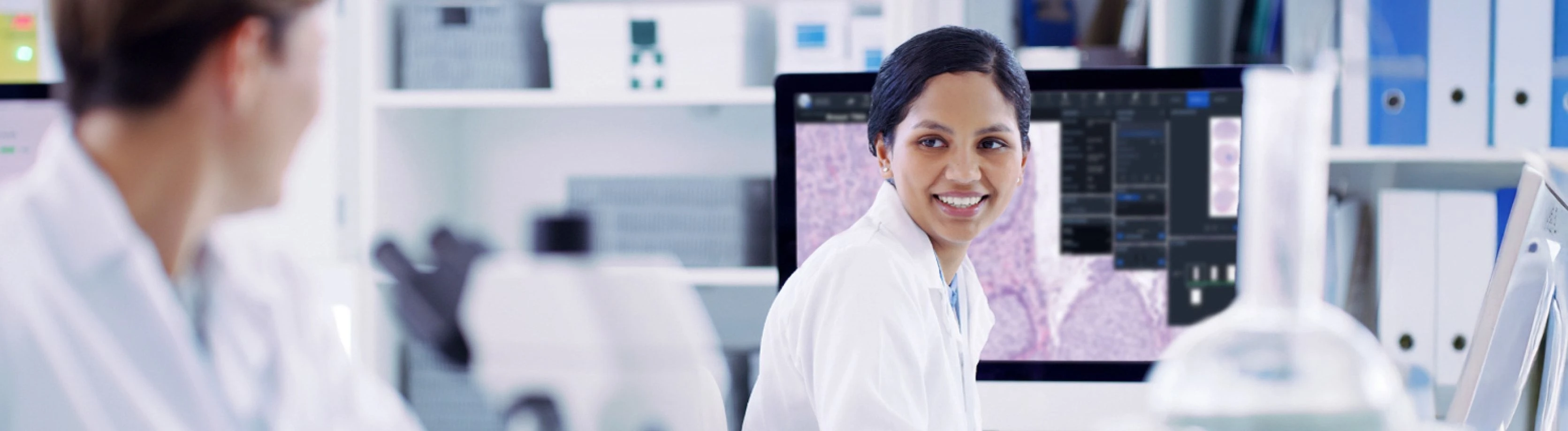 Two healthcare professionals in a laboratory. A female scientist in focus is smiling at a colleague while working on a computer displaying microscopic images, with lab equipment in the foreground.