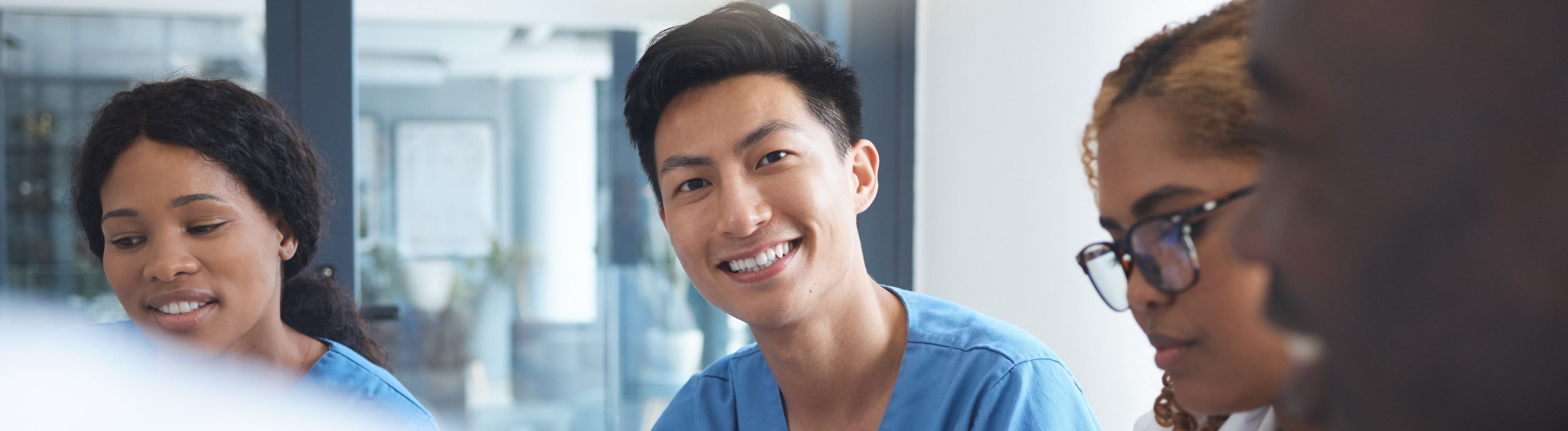 A smiling healthcare professional in scrubs during a collaborative meeting with colleagues in a bright office space.