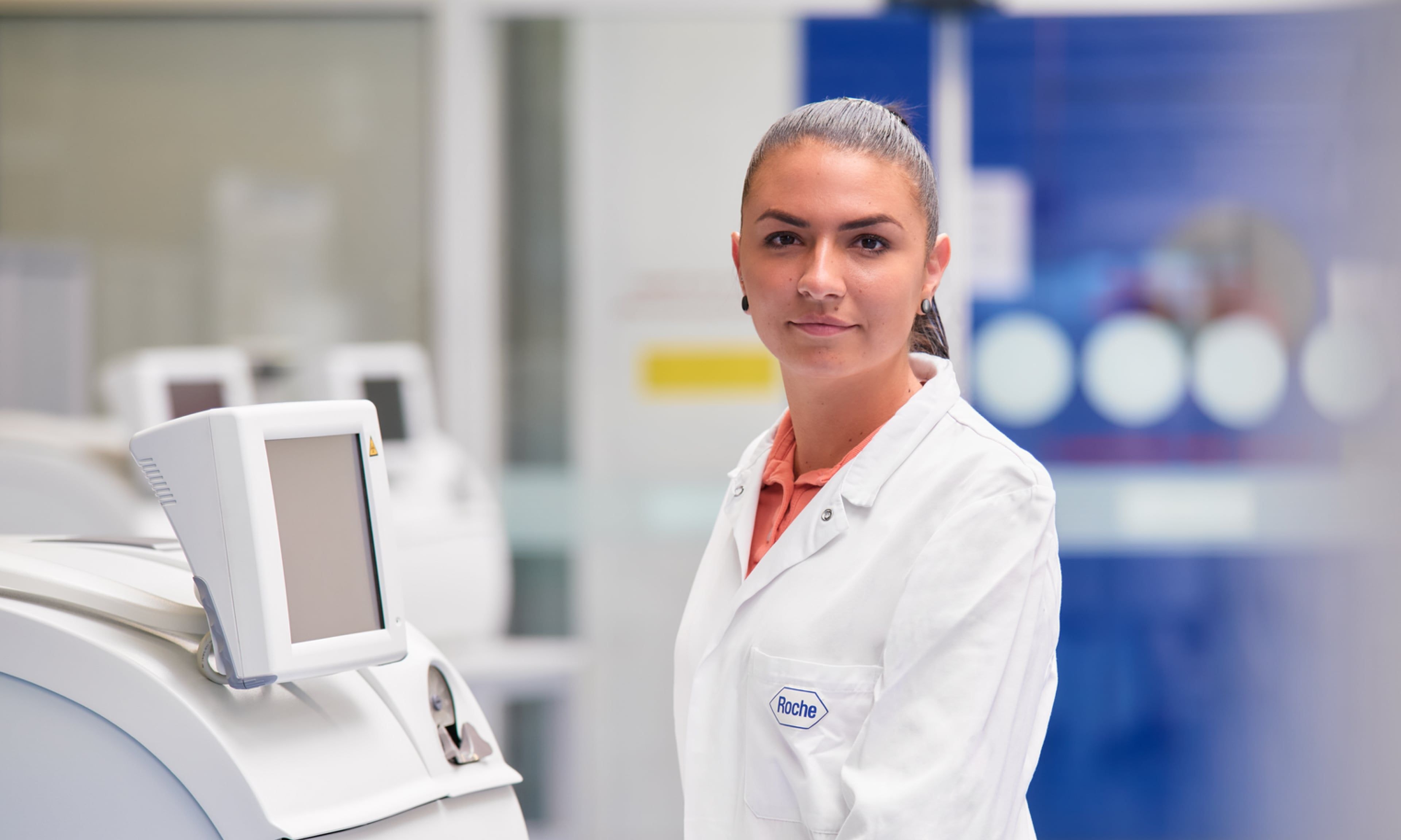 Female lab technician wearing a white coat standing by a monitor.