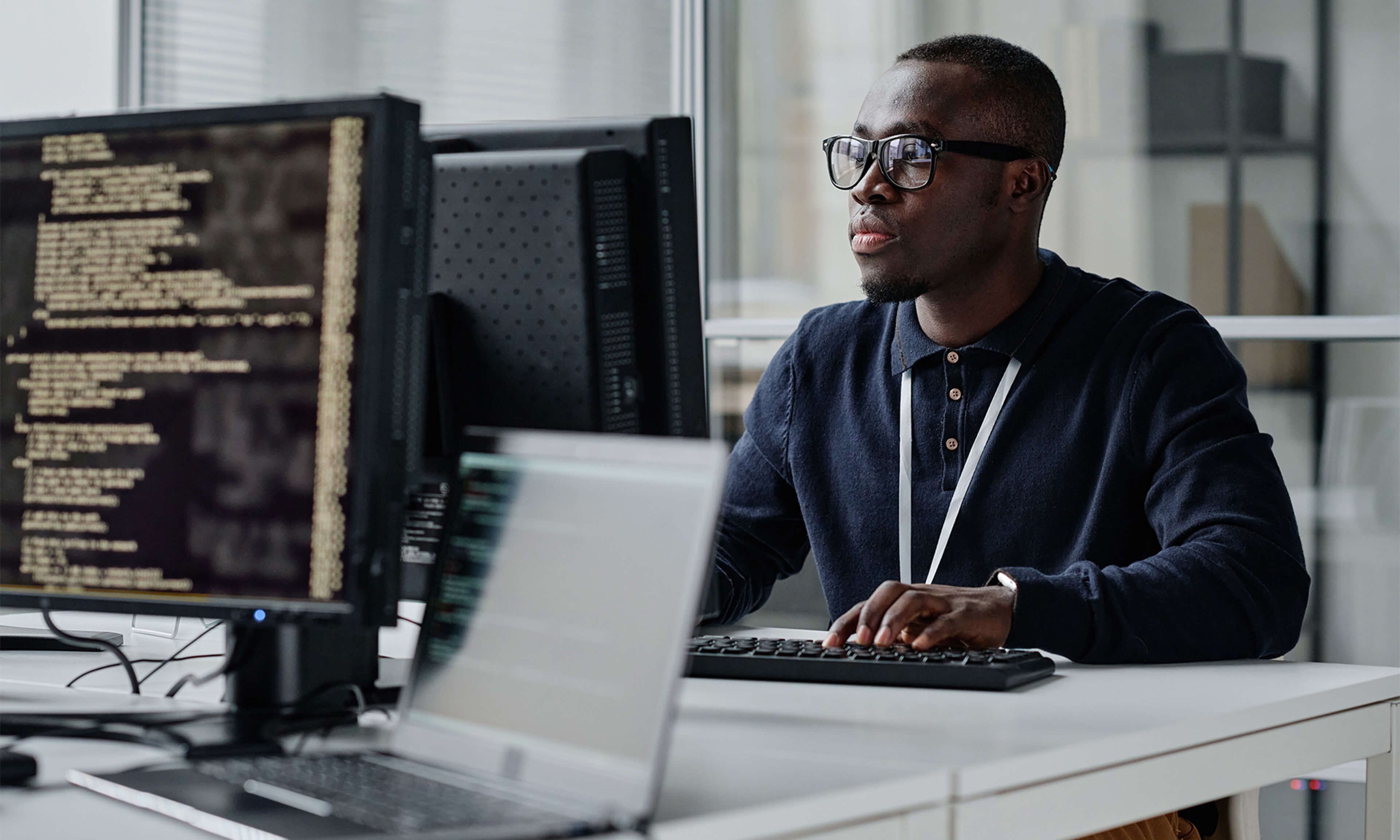 Male IT specialist focused on coding or system monitoring, working in a modern office with multiple monitors displaying data.