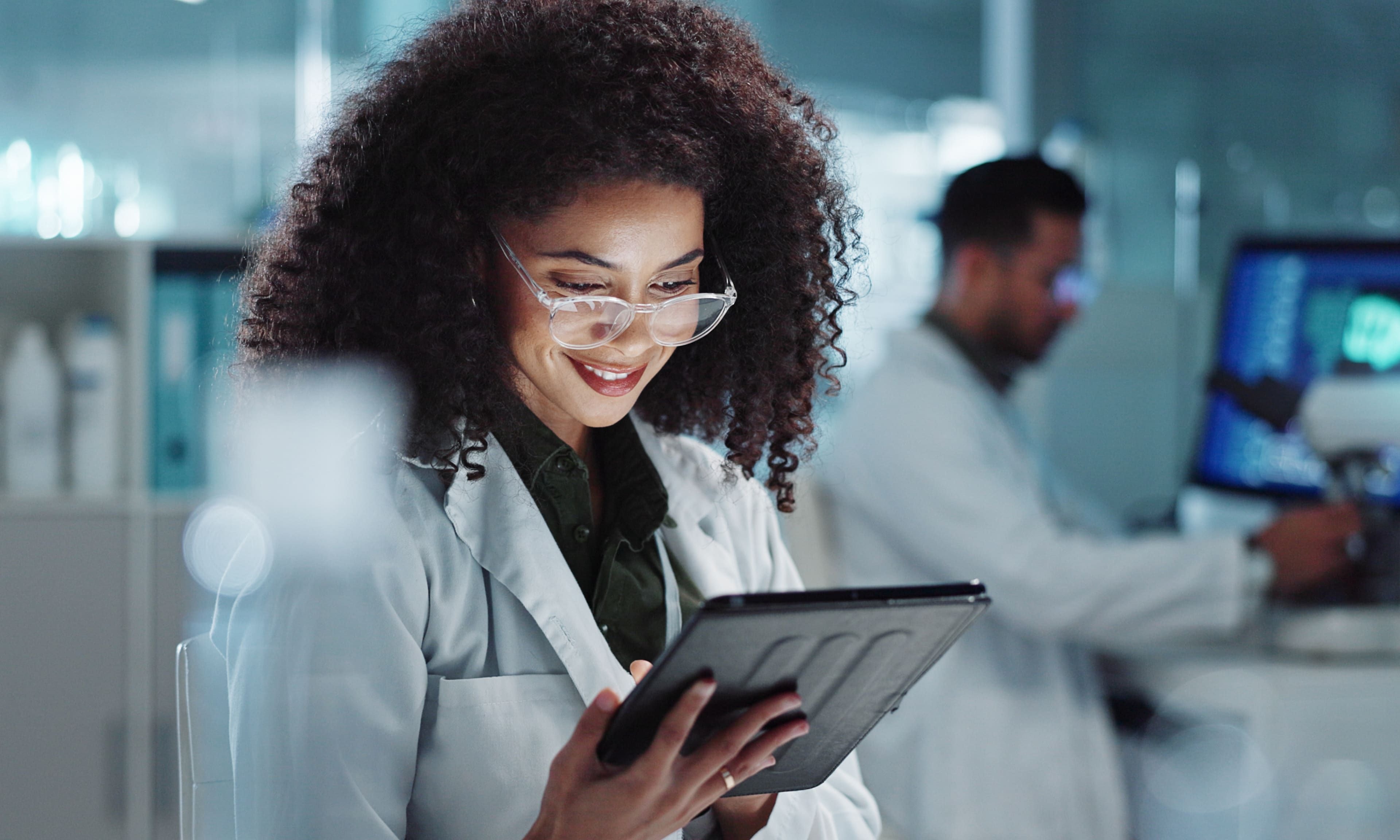 A smiling female scientist wearing glasses and a lab coat, working with a tablet in a modern laboratory. The background features another scientist at a computer station.