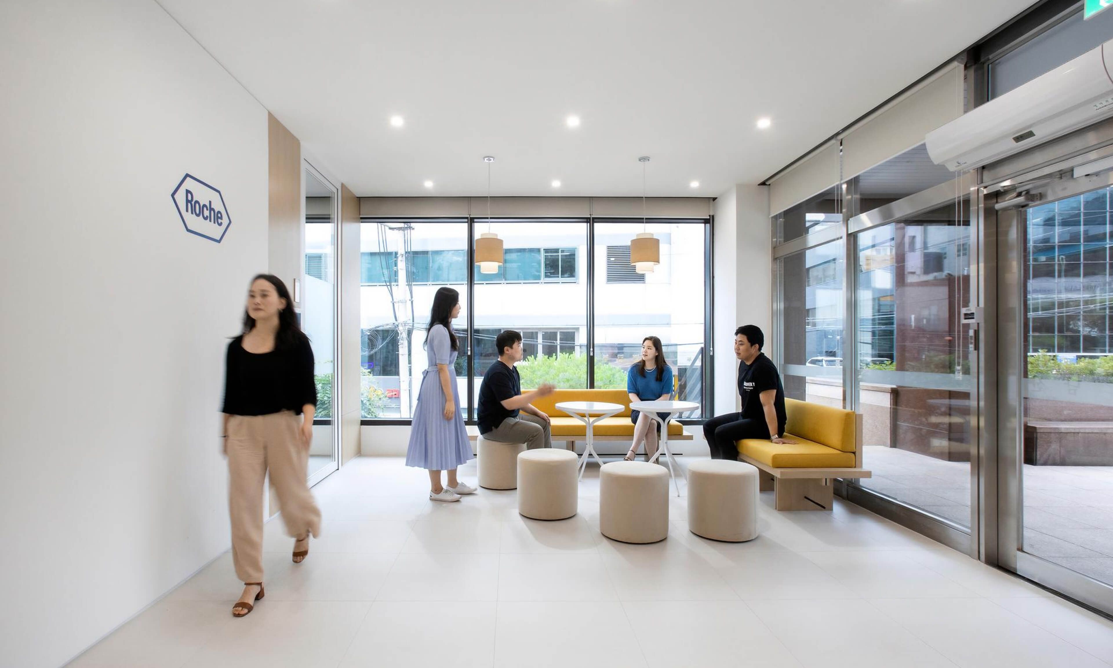 A modern office reception area with people interacting and walking. The space is bright, featuring large glass windows, yellow seating, and a Roche logo on the wall.
