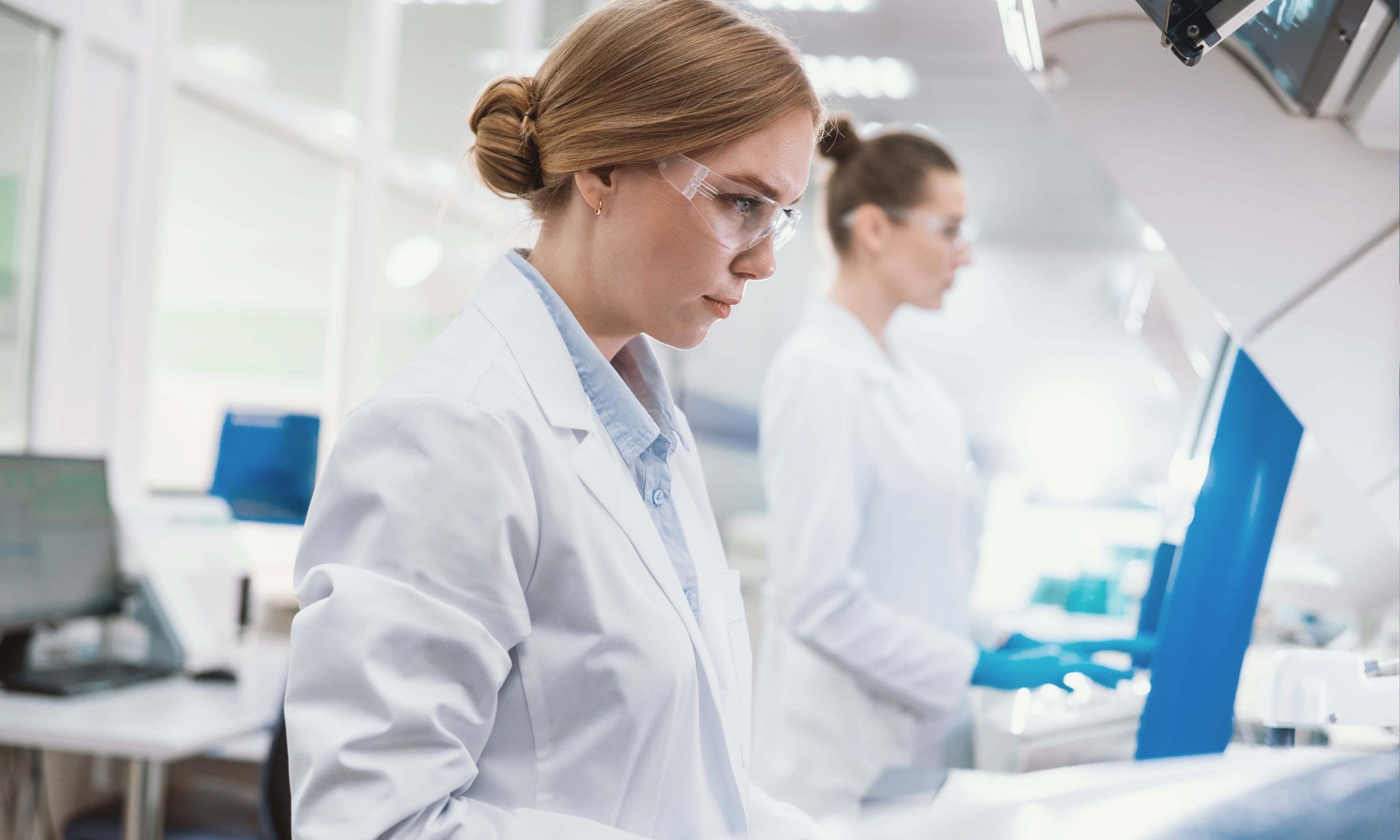 Female scientists in white lab coats and protective eyewear working with laboratory equipment in a bright, modern lab setting, focused on diagnostics or research.