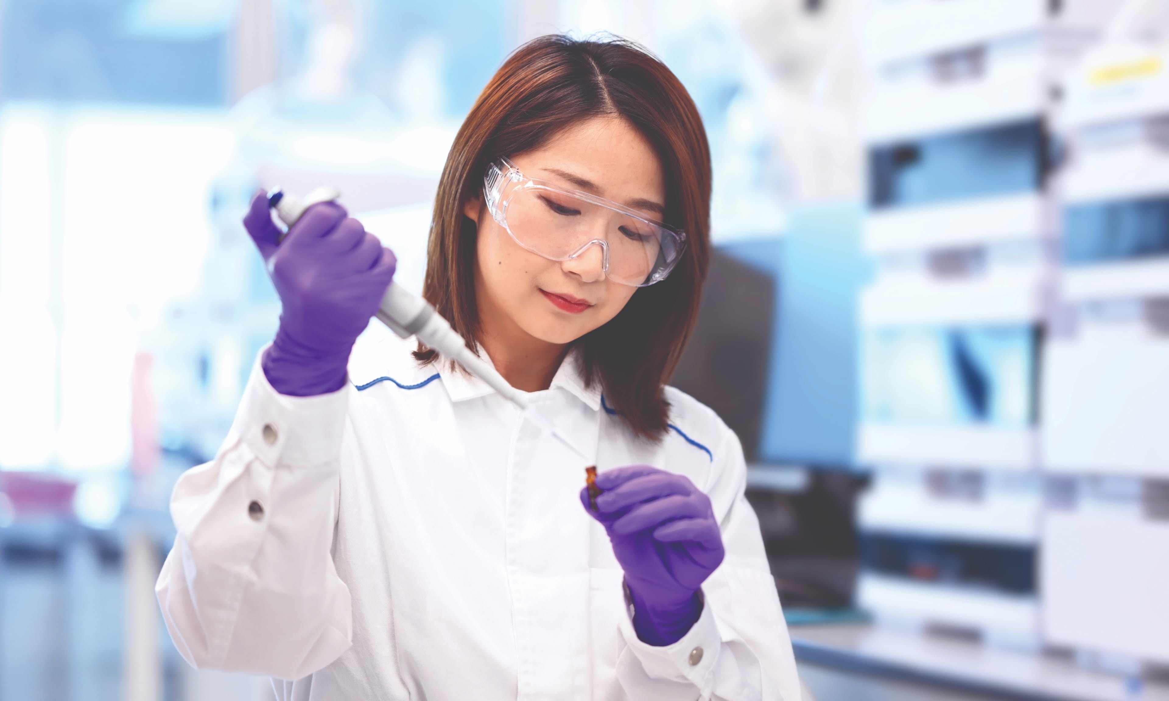 A scientist in a laboratory uses a pipette while wearing a white coat and purple gloves.