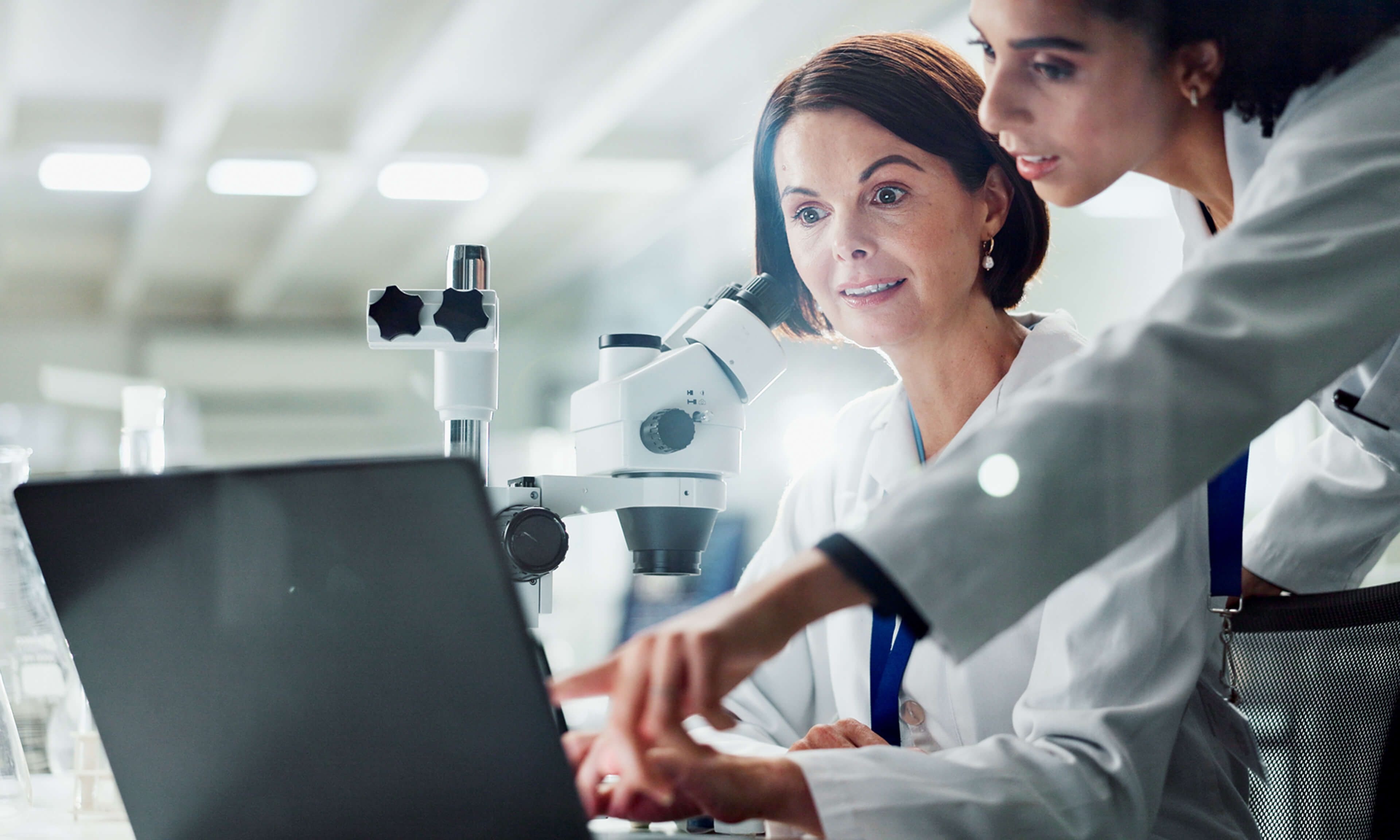 Deux femmes scientifiques dans un laboratoire discutant de données sur un ordinateur portable, avec un microscope et un équipement de laboratoire visibles en arrière-plan.