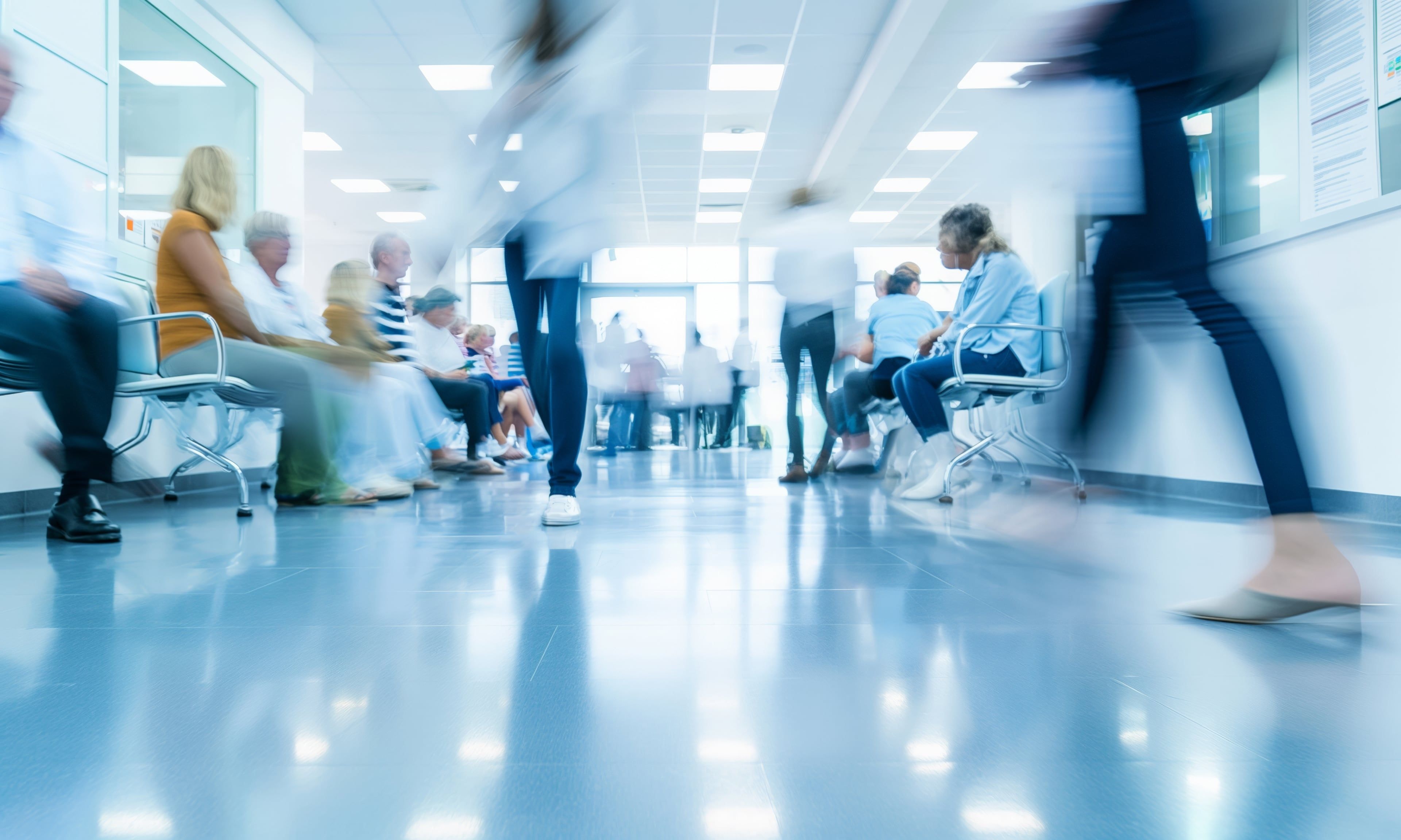 Blurred motion image of people walking through a hospital corridor with patients seated in a busy waiting area, indicating a fast-paced healthcare environment.